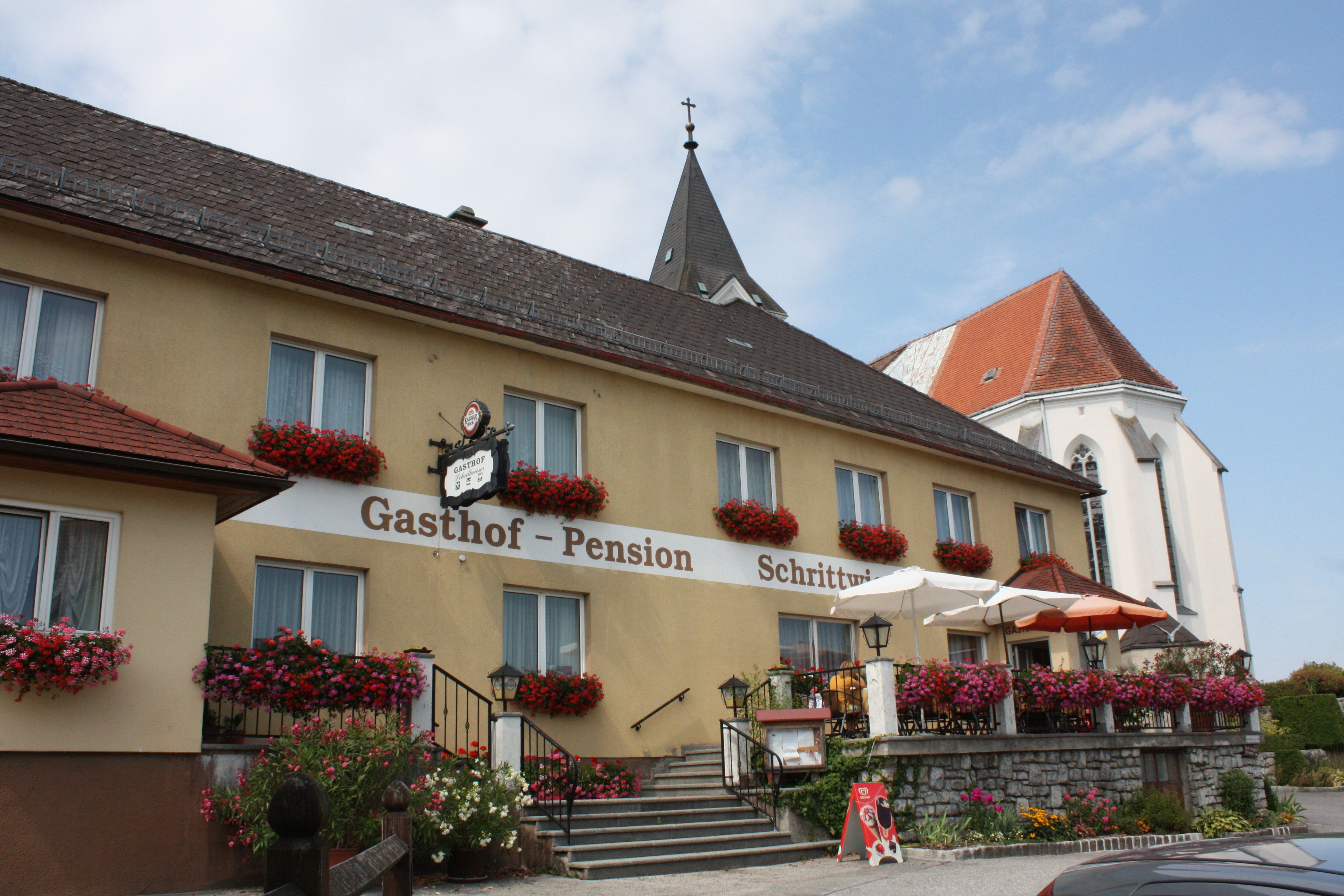 Ein traditioneller Gasthof mit Blumen geschmückter Fassade und Terrasse, daneben eine Kirche mit spitzem Turm.
