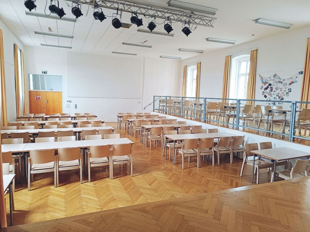 Ein leerer Saal mit Holzboden, Tischen und Stühlen, einer Bühne und Beleuchtung an der Decke.