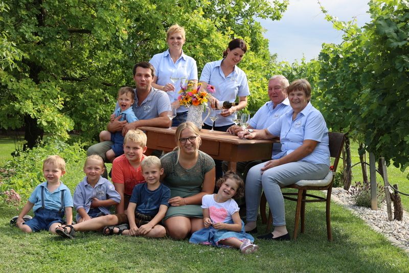 Eine Familie sitzt und steht um einen Tisch im Freien, umgeben von Bäumen und Gras.