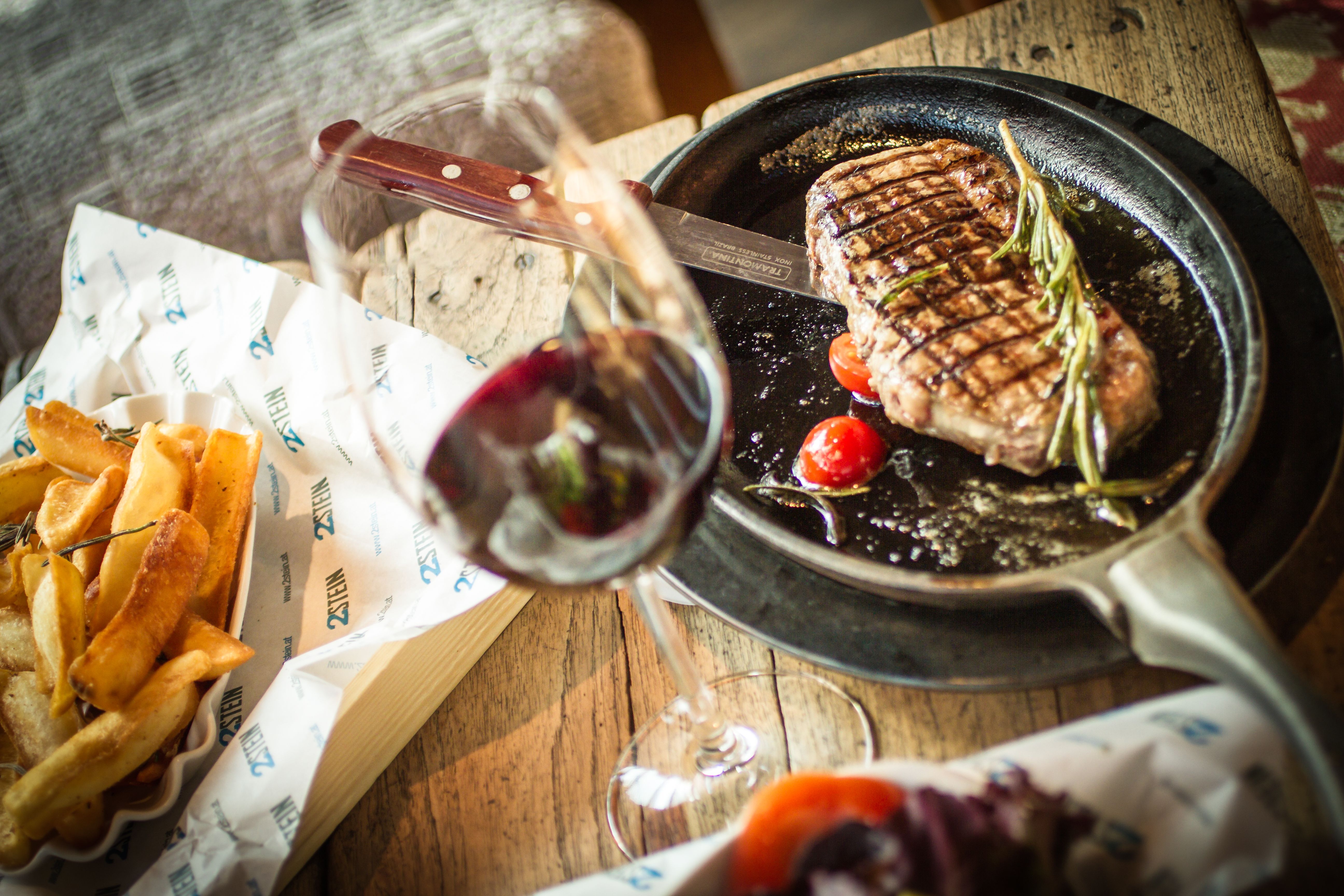 Ein Steak in einer Pfanne mit Tomaten und Rosmarin, daneben Pommes und ein Glas Rotwein auf einem Holztisch.