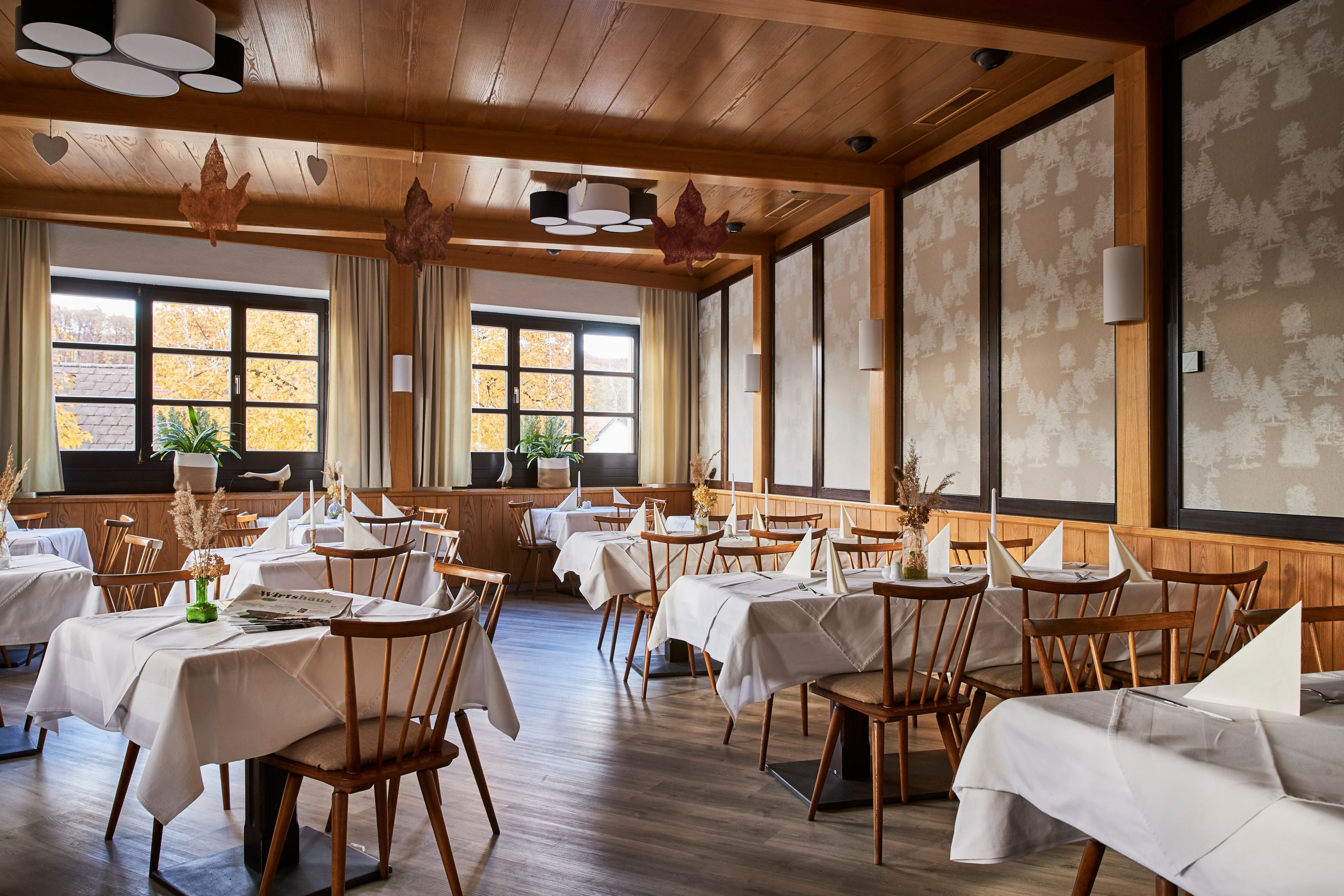Ein elegantes Restaurant mit Holzmöbeln, weißen Tischdecken und großen Fenstern, die herbstliche Bäume zeigen.
