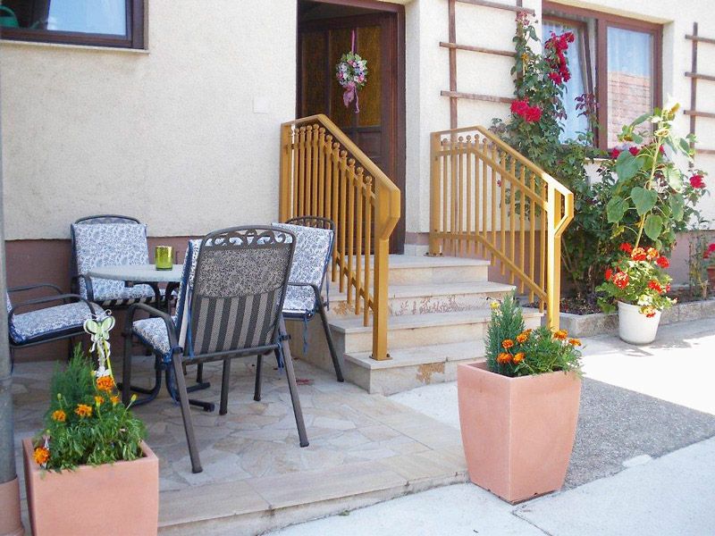 Eingangsbereich einer Ferienwohnung mit Terrasse, Tisch und Stühlen, Blumen und Geländer.