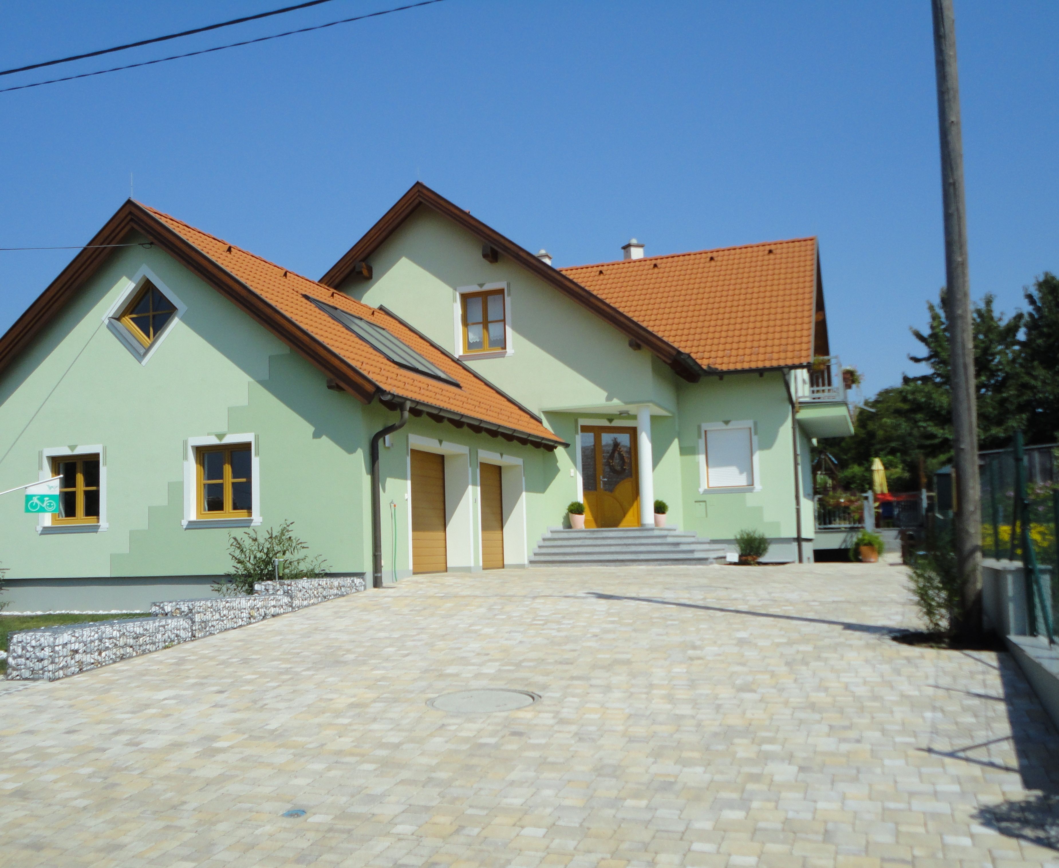 Ein modernes, zweistöckiges Haus mit grünem Anstrich und roten Dachziegeln, umgeben von einem gepflasterten Hof.