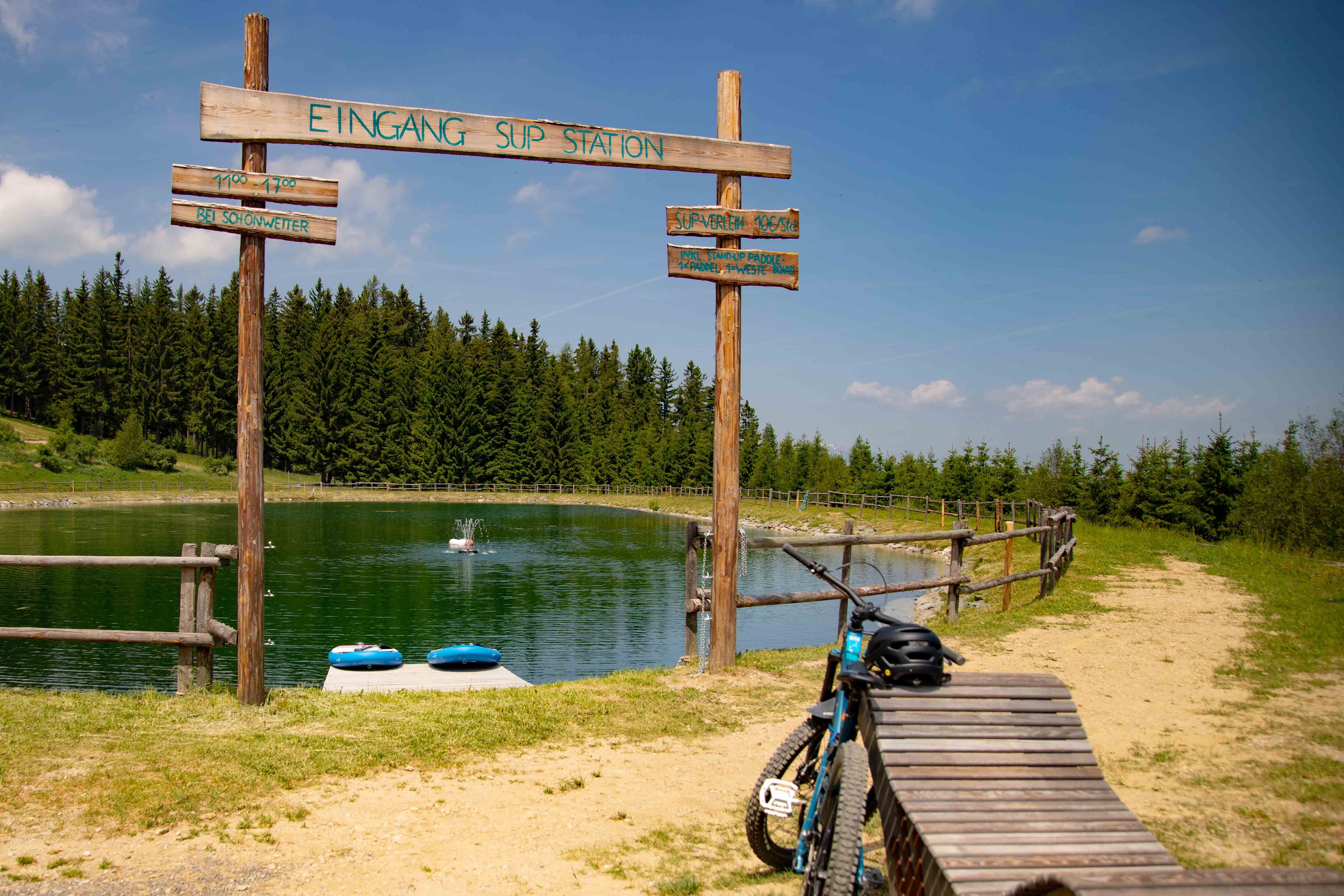 Speicherteich mit SUP-Station in St. Corona am Wechsel, umgeben von Wald und einem Fahrrad im Vordergrund.