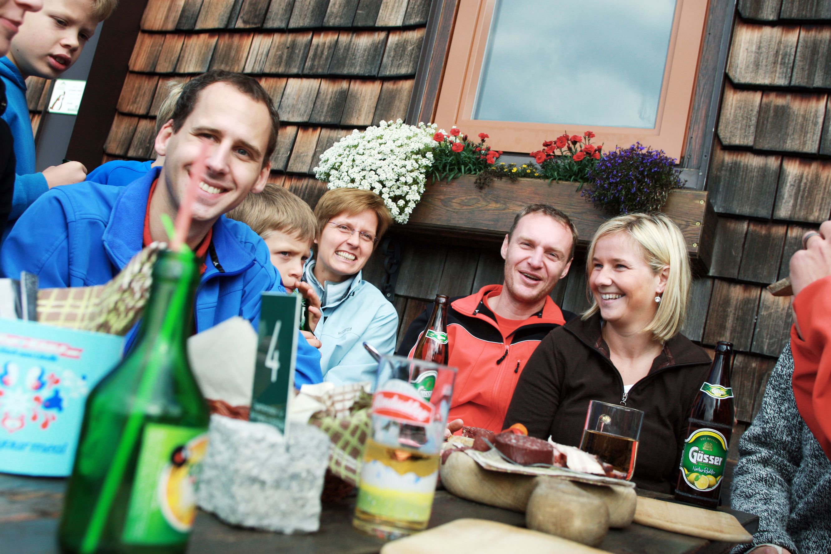 Gruppe von Menschen sitzt lachend an einem Tisch vor einer Holzhütte mit Blumen.