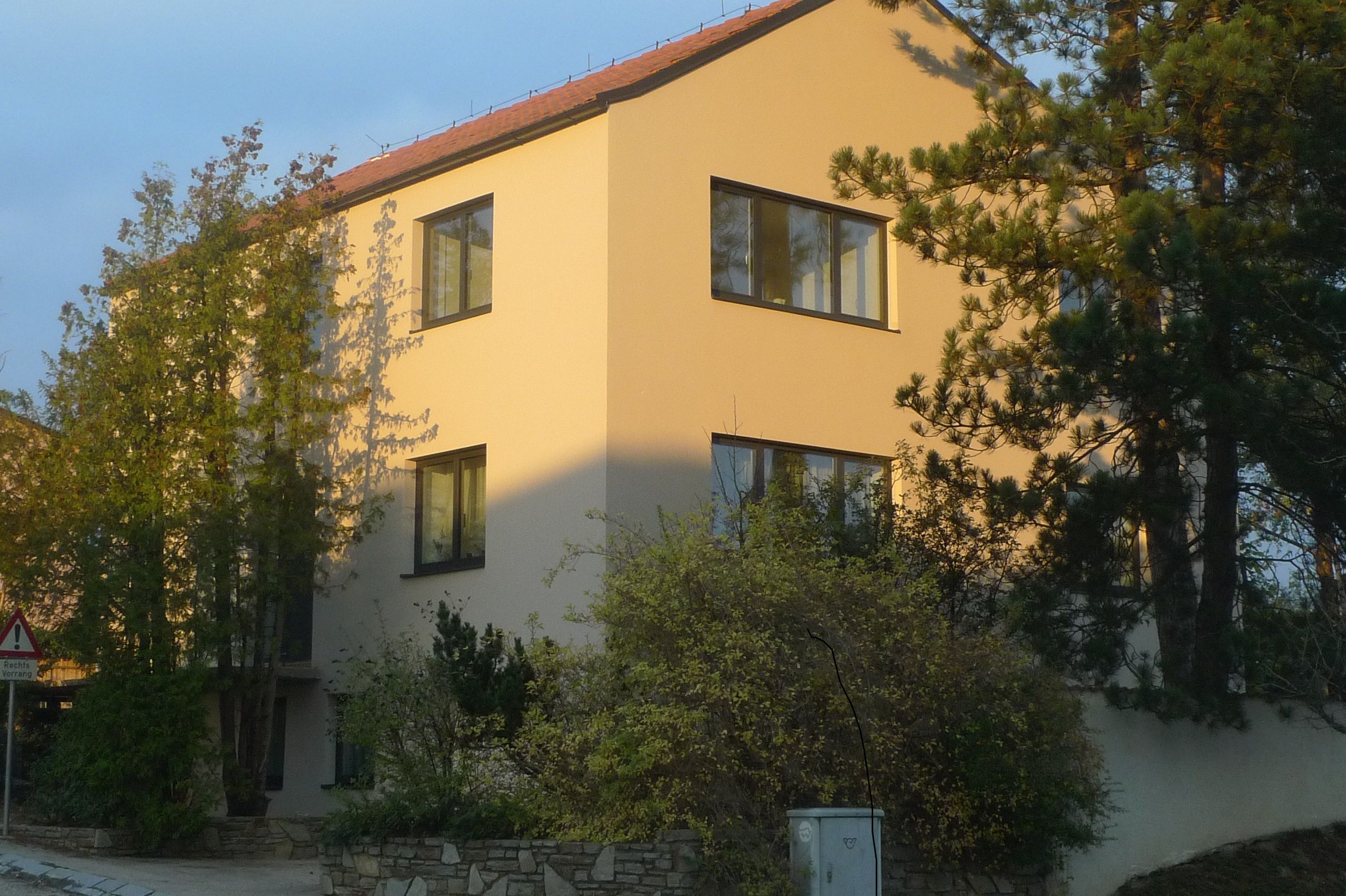 Ein modernes, dreistöckiges Haus mit gelber Fassade und roten Dachziegeln, umgeben von Bäumen und Sträuchern.