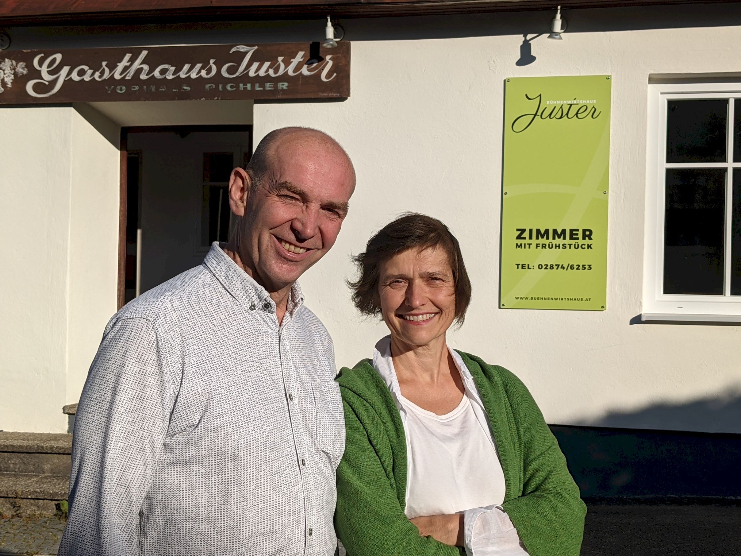 Ein Mann und eine Frau stehen lächelnd vor dem Gasthaus Jüster. Im Hintergrund ist ein Schild mit der Aufschrift 'Zimmer mit Frühstück' zu sehen.