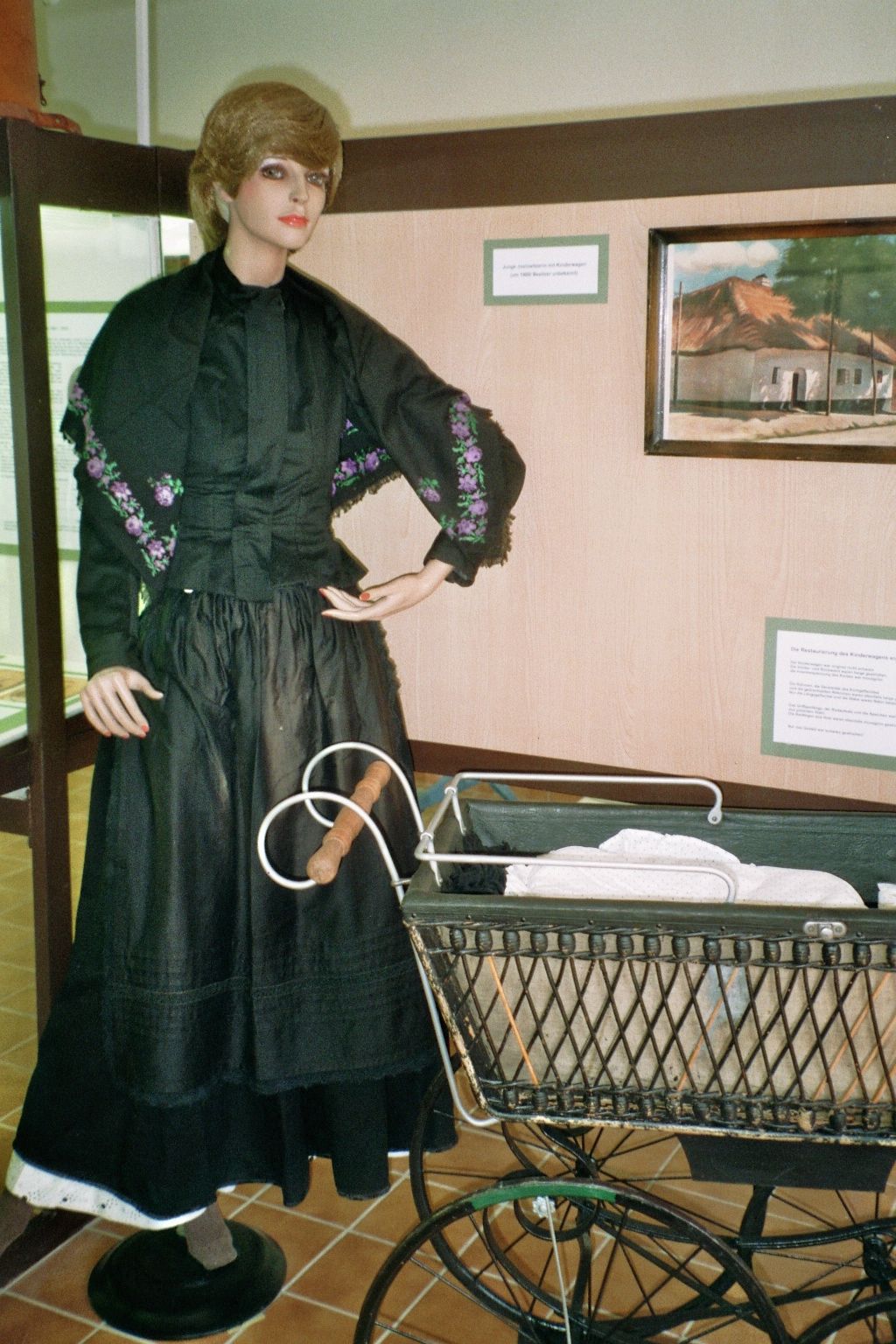 Eine Schaufensterpuppe in traditioneller Kleidung neben einem alten Kinderwagen in einem Ausstellungsraum.