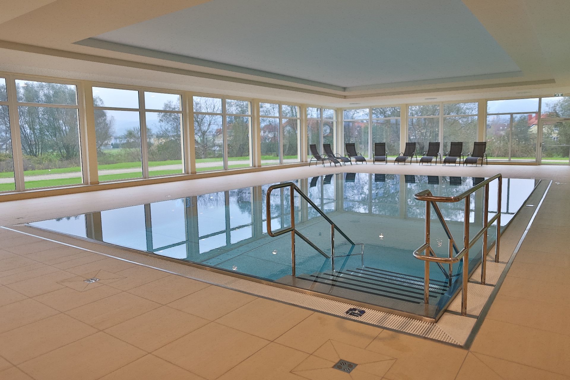 Innenansicht eines Hallenbads mit großen Fenstern und Liegestühlen am Beckenrand.