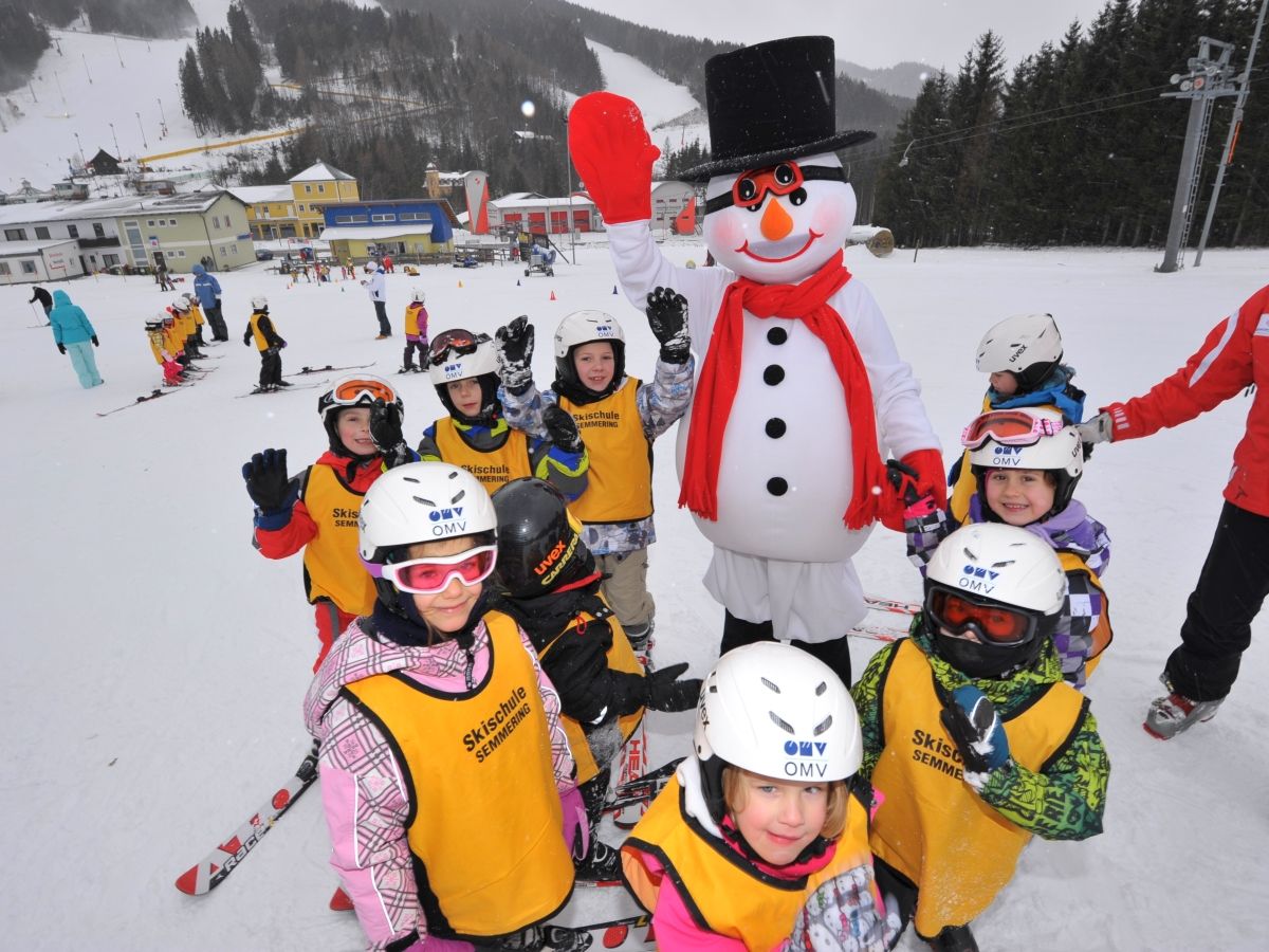 Kinder in Skiausrüstung posieren mit einem Schneemann auf einer Skipiste.