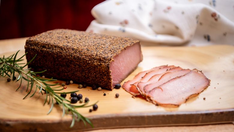 Ein Stück gepökeltes Fleisch mit Pfefferkruste und geschnittenen Scheiben auf einem Holzbrett, dekoriert mit Rosmarin und Wacholderbeeren.