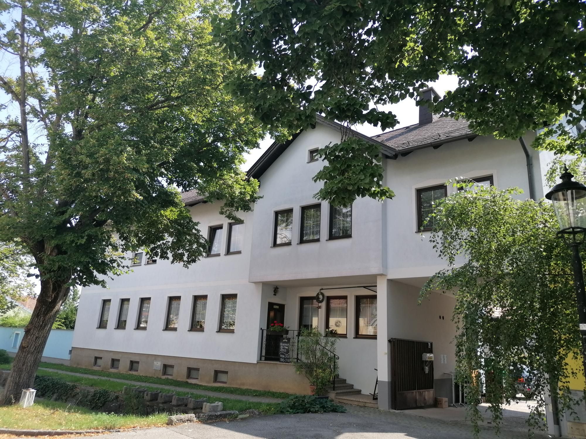 Ein weißes Gebäude mit mehreren Fenstern, umgeben von Bäumen und einem Laternenpfahl.