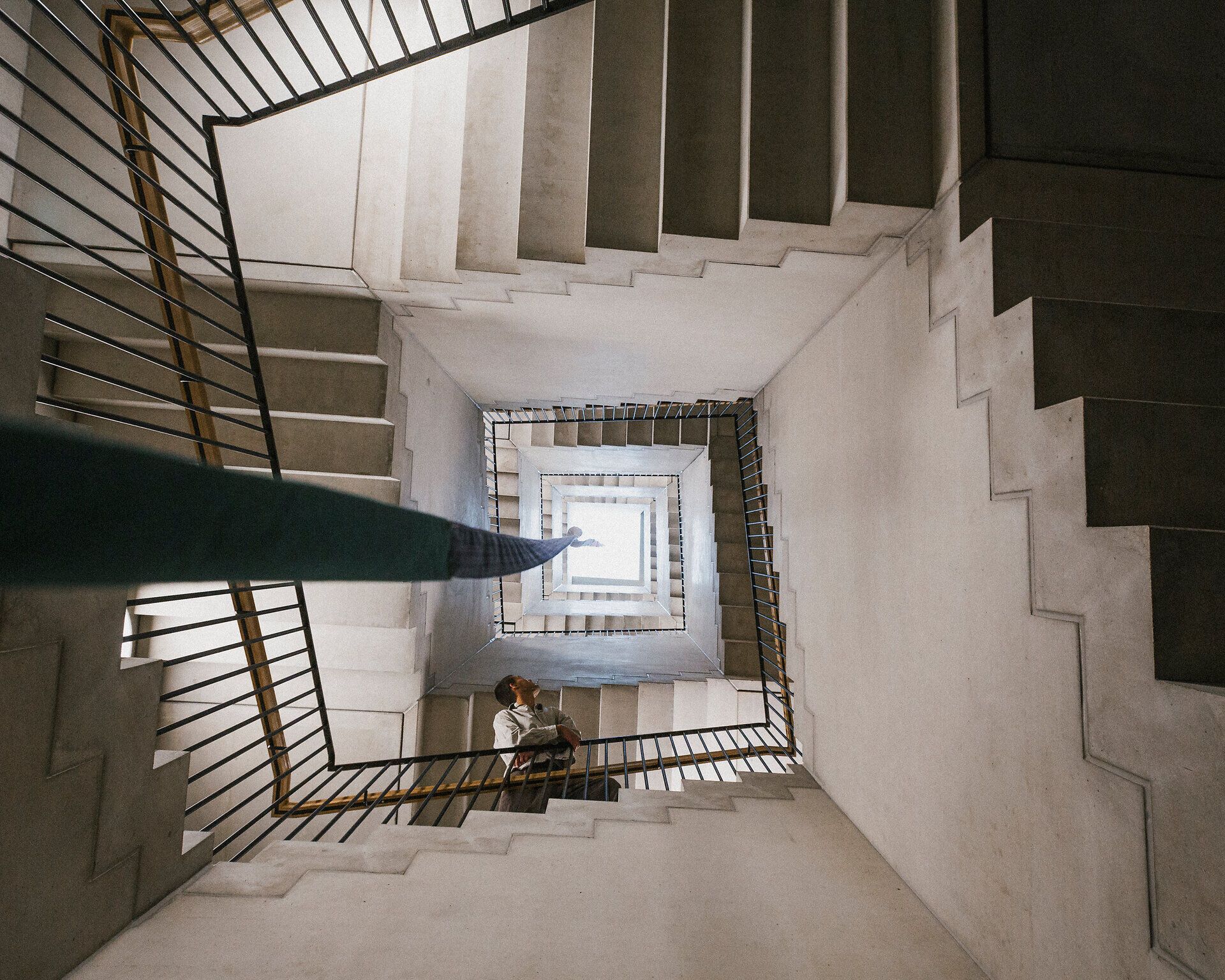 Blick von oben in ein quadratisches Stiegenhaus mit mehreren Treppenläufen und Metallgeländern; unten ist eine Person auf dem Zwischenpodest zu sehen.