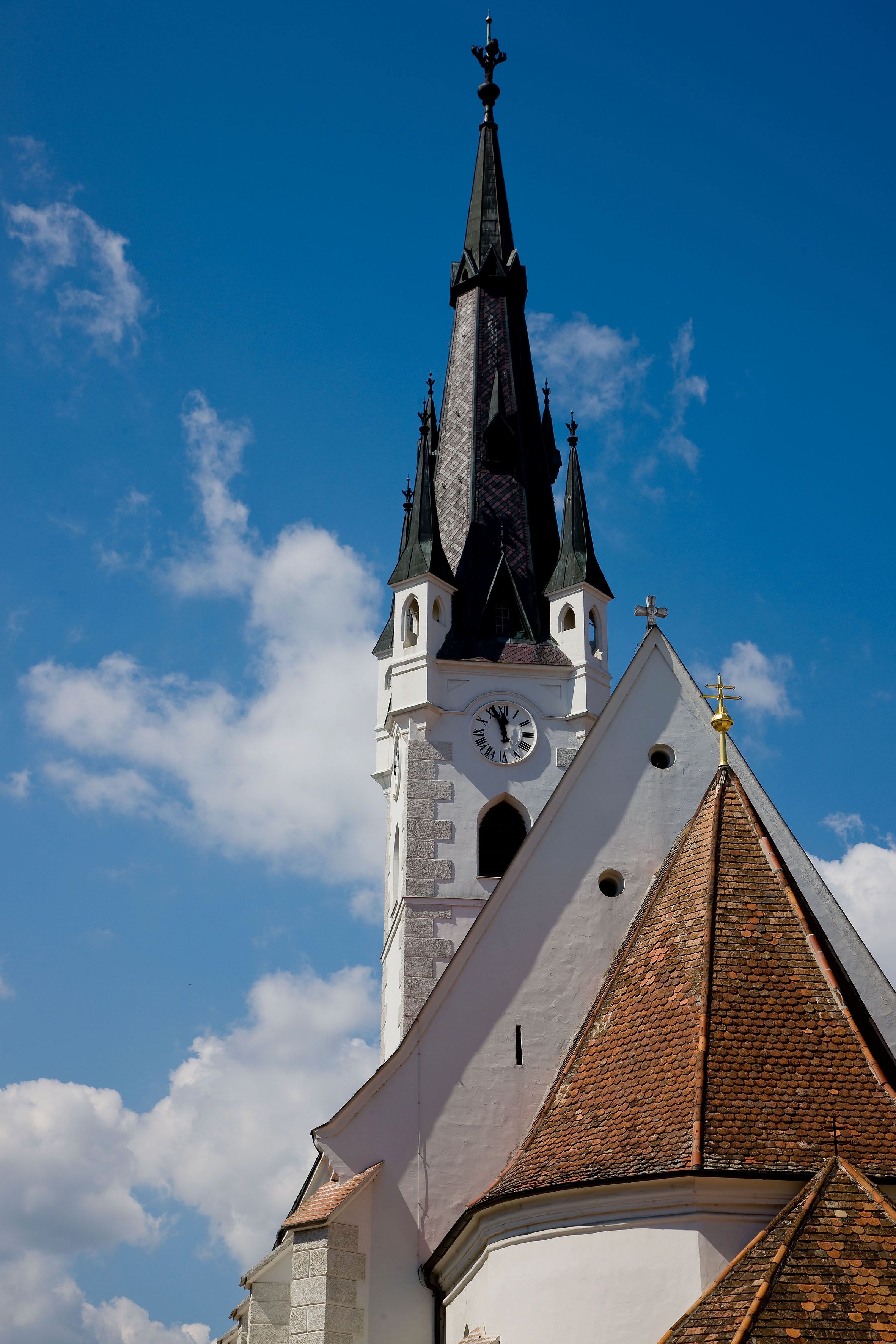 Kirchturm mit Uhr und spitzem Dach vor blauem Himmel.