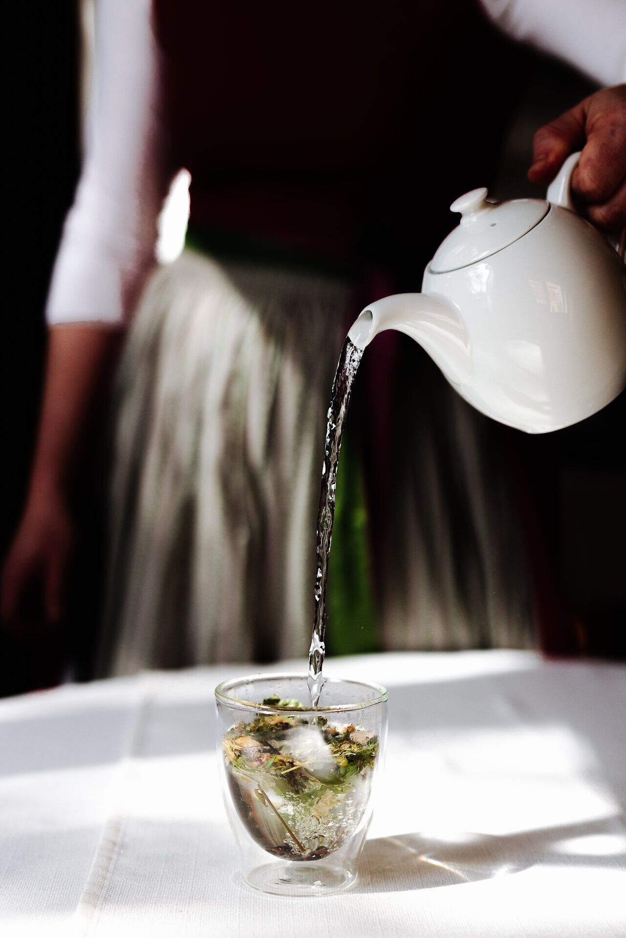 Ein zarter Wasserstrahl fließt in ein Glas, das mit duftenden Wildkräutern gefüllt ist. Die sanften Lichtstrahlen schaffen eine einladende Atmosphäre, die zum Genießen und Entspannen einlädt. Hier wird die Essenz der Natur in einer Tasse Tee lebendig.