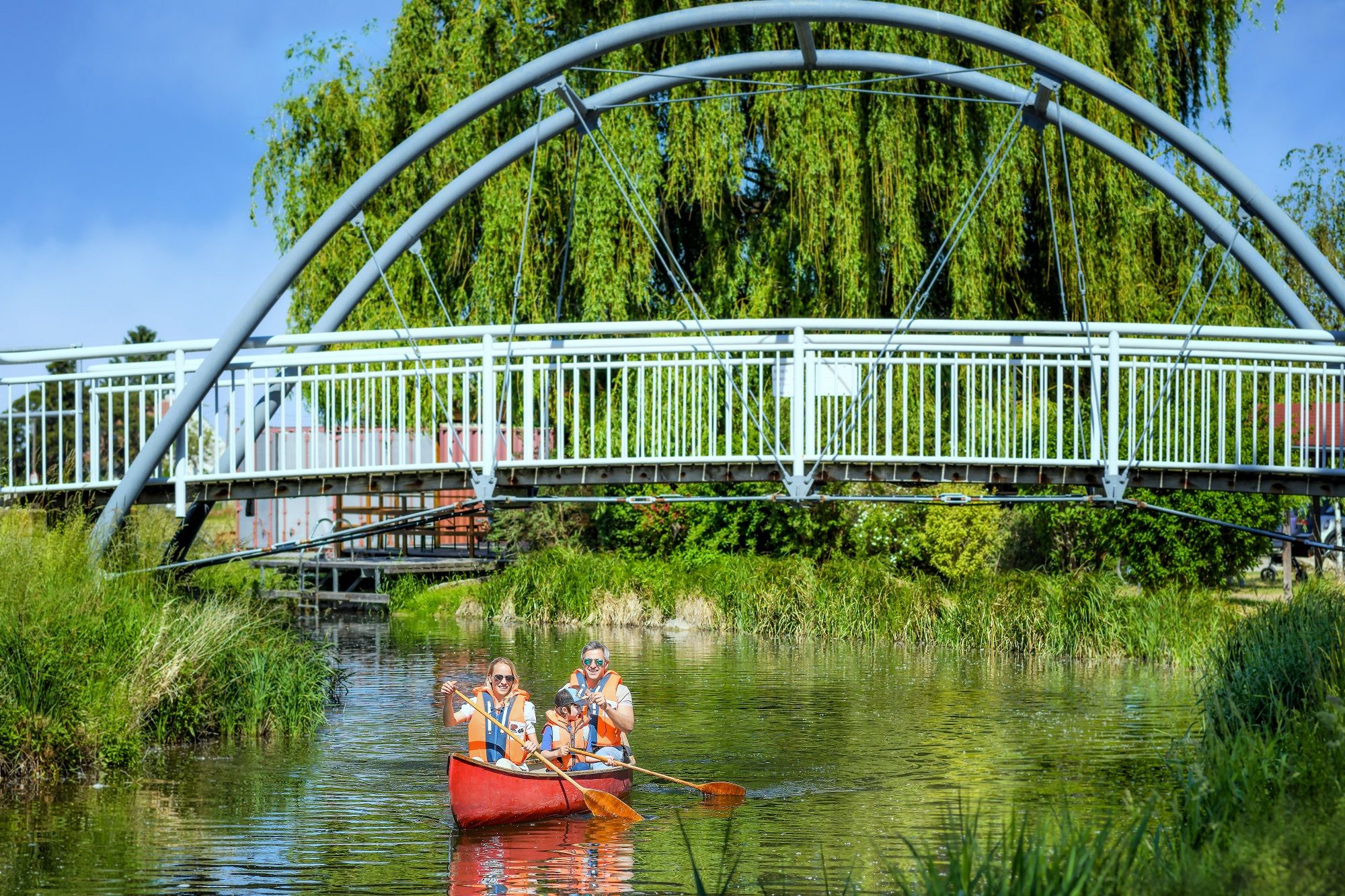 Schifferlfahren am Wiener Neustädter Kanal, ein rotes Boot mit einer Familie fährt unter einer Brücke am Kanal durchs Grüne