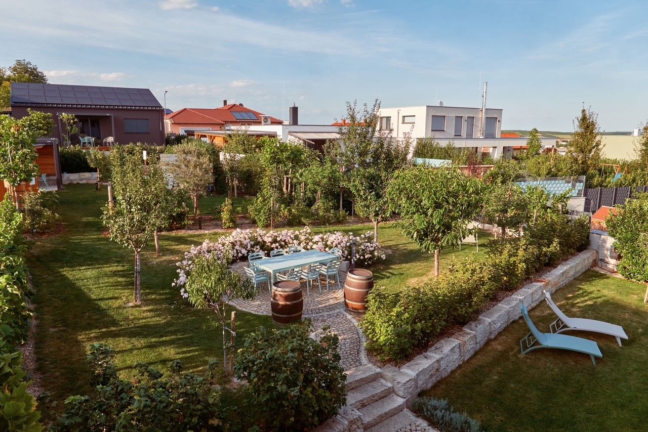 Ein gepflegter Garten mit Bäumen, Blumen, einem Tisch mit Stühlen und zwei Liegestühlen. Im Hintergrund sind moderne Häuser zu sehen.