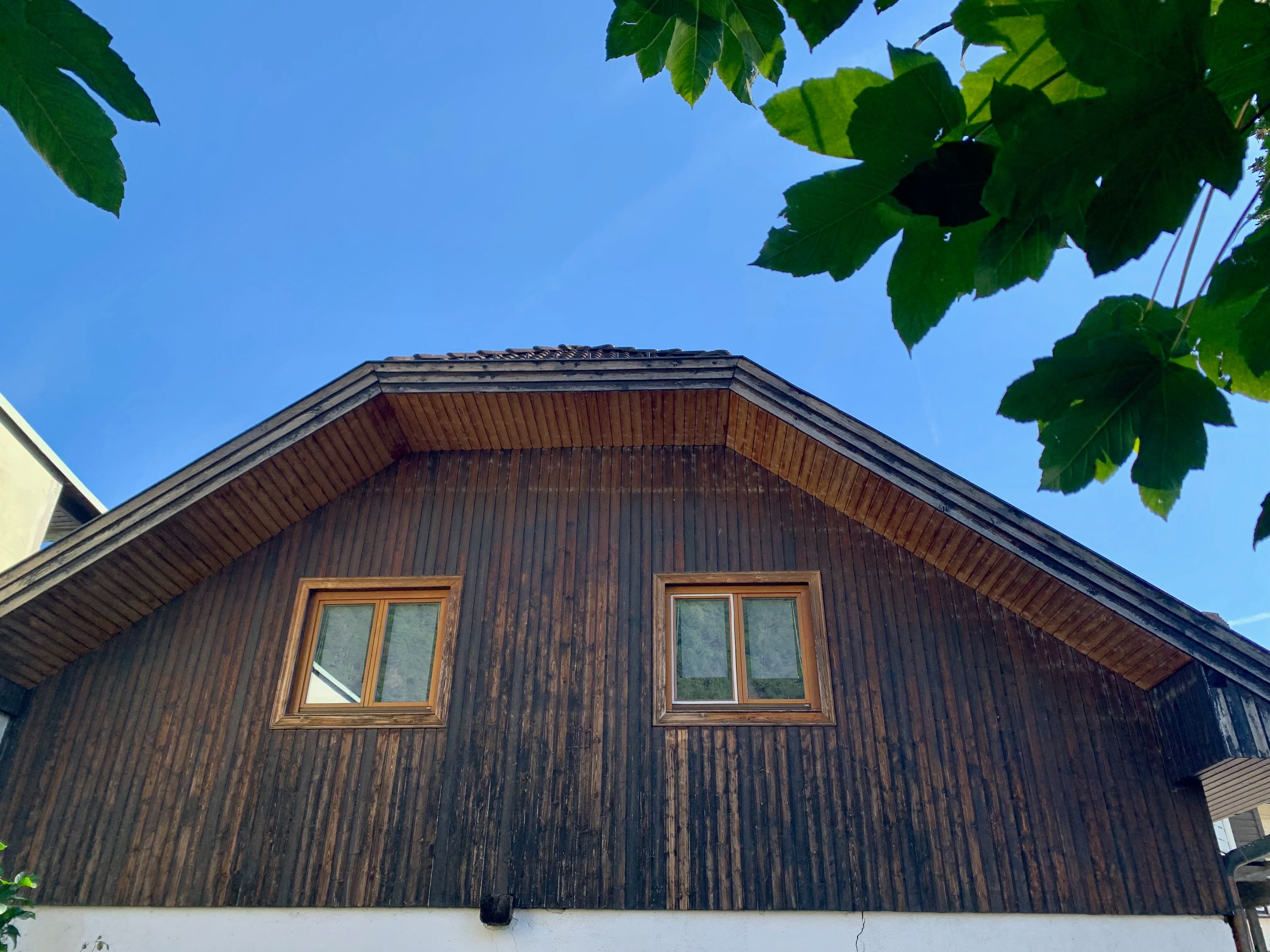 Dachgeschoss eines Hauses mit Holzverkleidung und zwei Fenstern, umgeben von grünen Blättern und blauem Himmel.