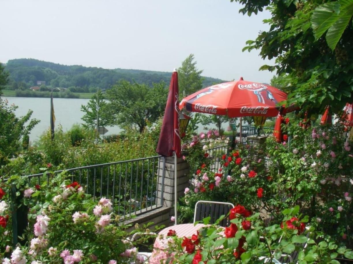 Ein blühender Garten mit roten und rosa Rosen, einem Tisch mit Stühlen und einem roten Sonnenschirm mit Coca-Cola-Logo, im Hintergrund ein See.