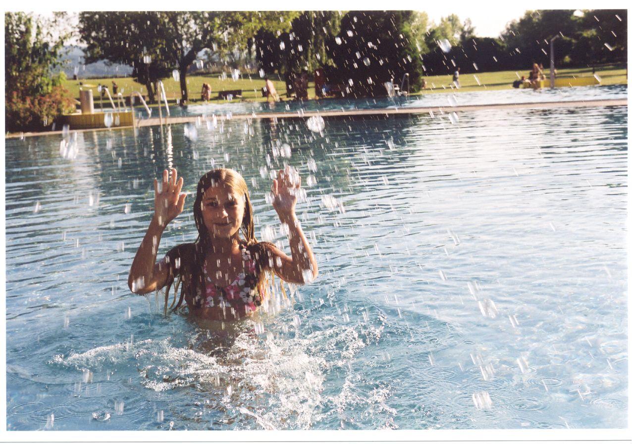 Ein Mädchen im Wasser eines Freibads, umgeben von spritzendem Wasser, mit Bäumen und Wiese im Hintergrund.