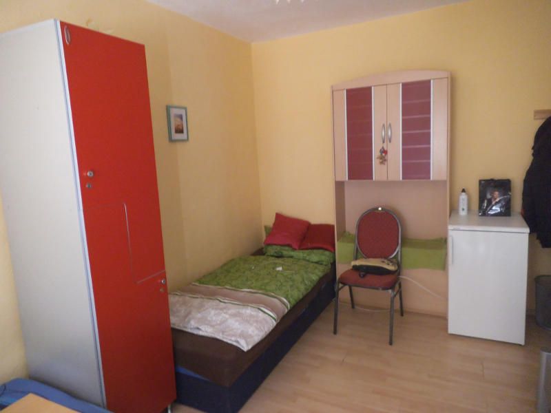Ein kleines Einzelzimmer mit Bett, rotem Schrank, Stuhl und kleinem Kühlschrank.