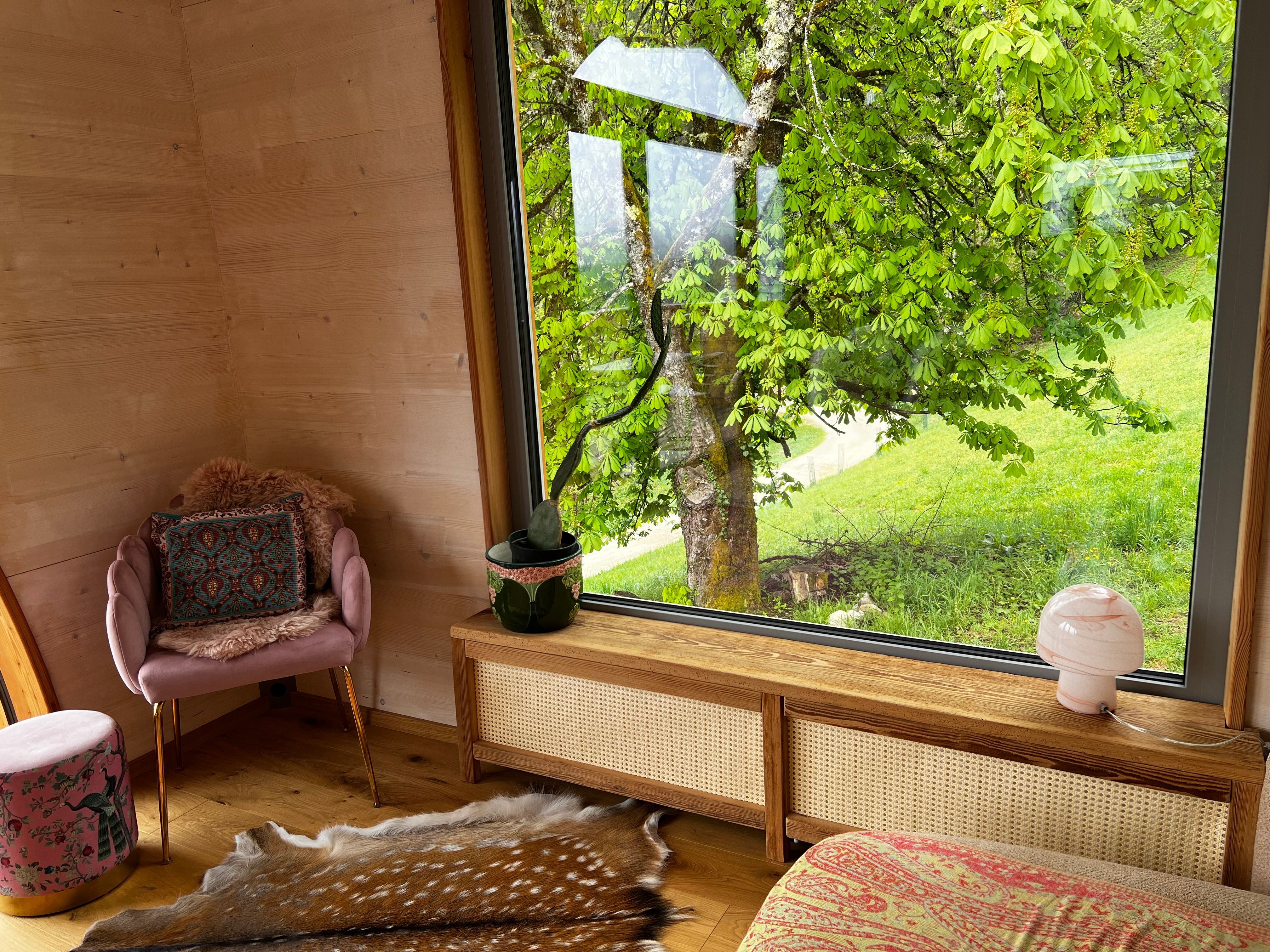 Innenraum eines gemütlichen Zimmers mit großem Fenster und Blick auf einen Baum und grüne Wiese.