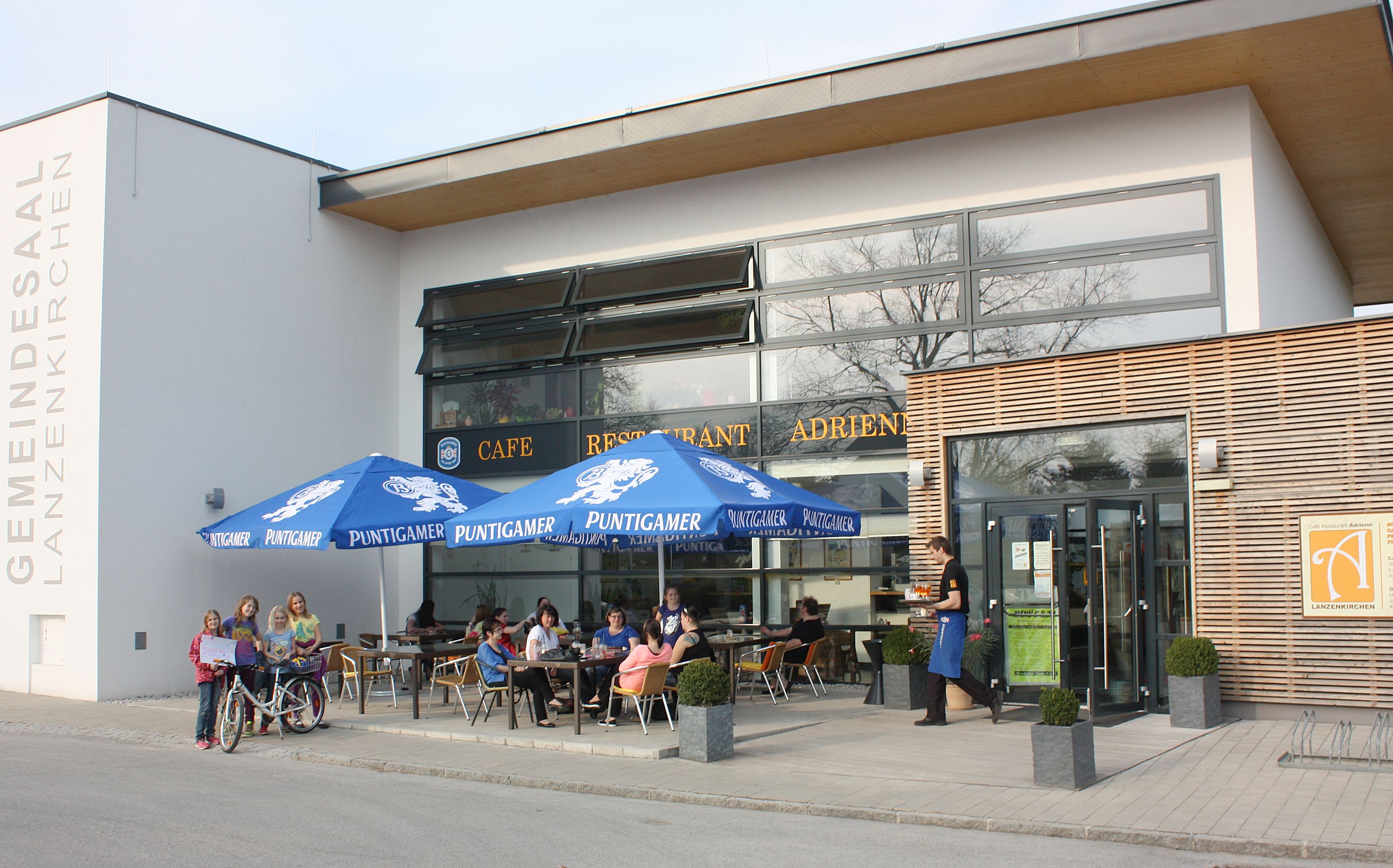 Außenansicht des Cafe Adrienn mit Gästen unter blauen Sonnenschirmen.