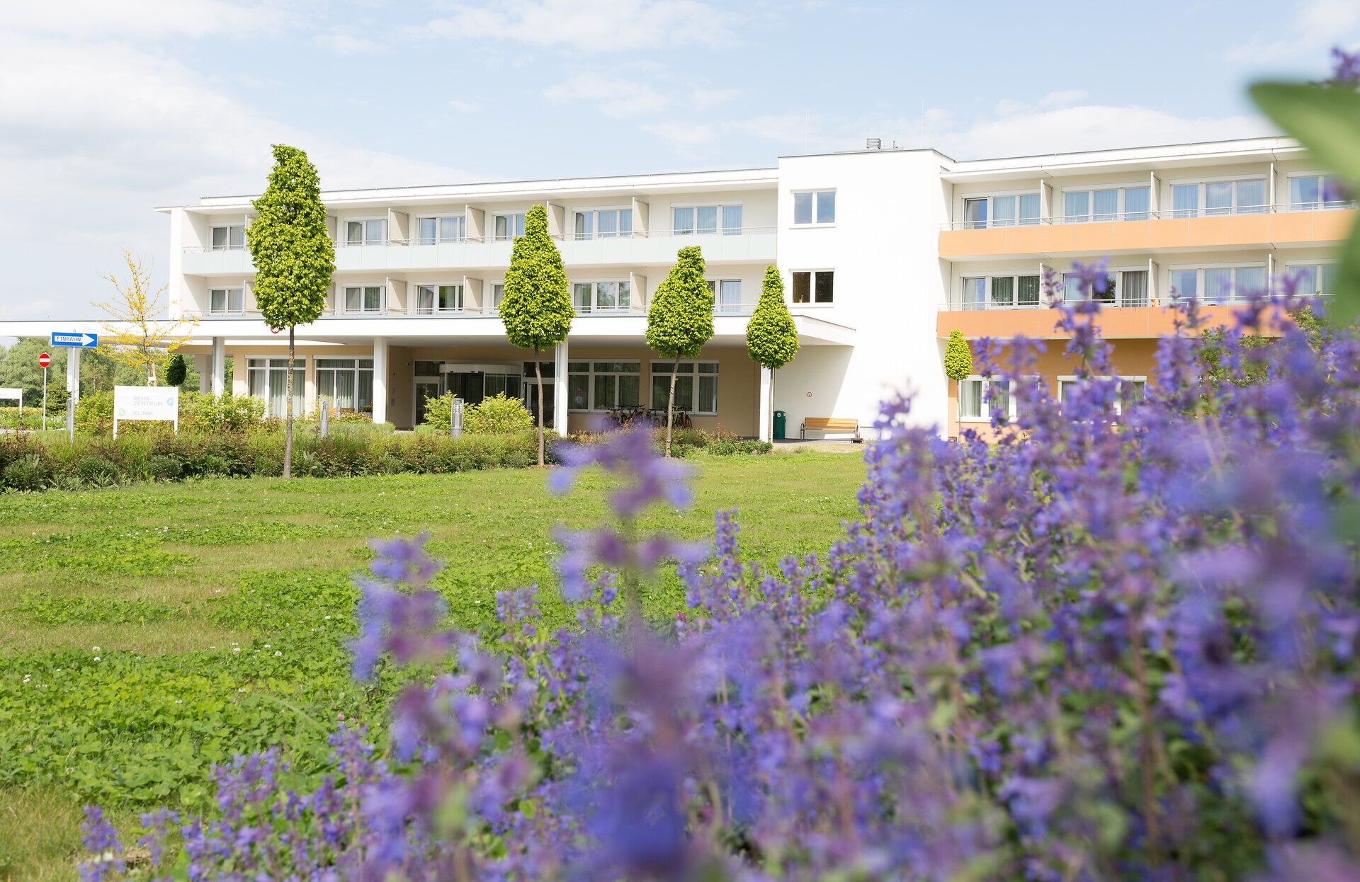 Außenansicht eines dreistöckigen Gebäudes mit vielen Fenstern und Balkonen und Blick auf den überdachten Eingangsbereich. Im Vordergrund ist eine Gartenanlage zu erkennen.