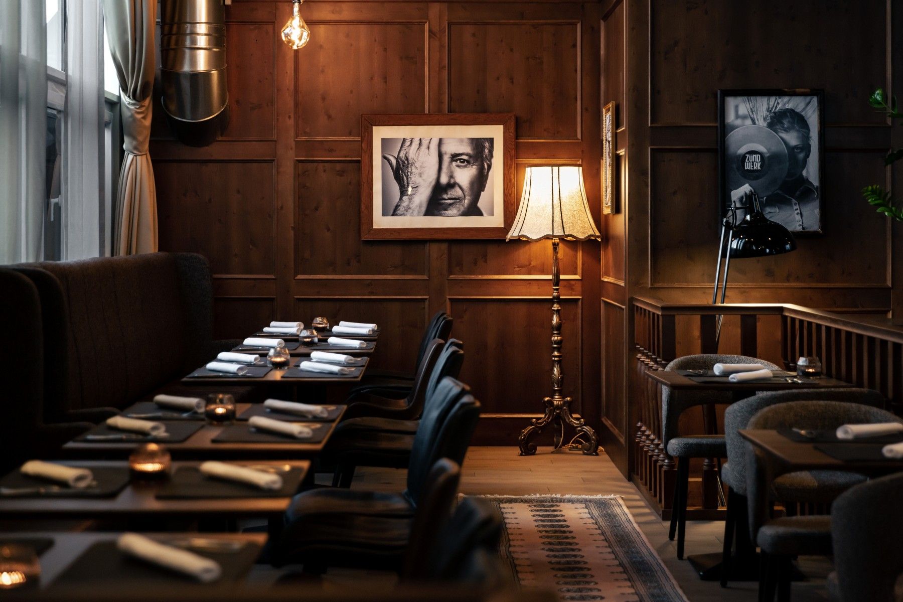 Gemütlicher Raum mit Holzwänden, gedeckten Tischen, Stehlampe und Schwarz-Weiß-Fotografien an der Wand.