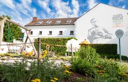 Ein einladender Spielplatz umgeben von blühenden Pflanzen und einer beeindruckenden Wandmalerei, die die Kreativität und den Geist der Kunst feiert. Hier können Familien die frische Luft genießen und die Schönheit der Natur erleben, während die Kinder fröhlich spielen. Ein Ort, der sowohl für Kunstliebhaber als auch für Naturliebhaber ein wahres Paradies ist.