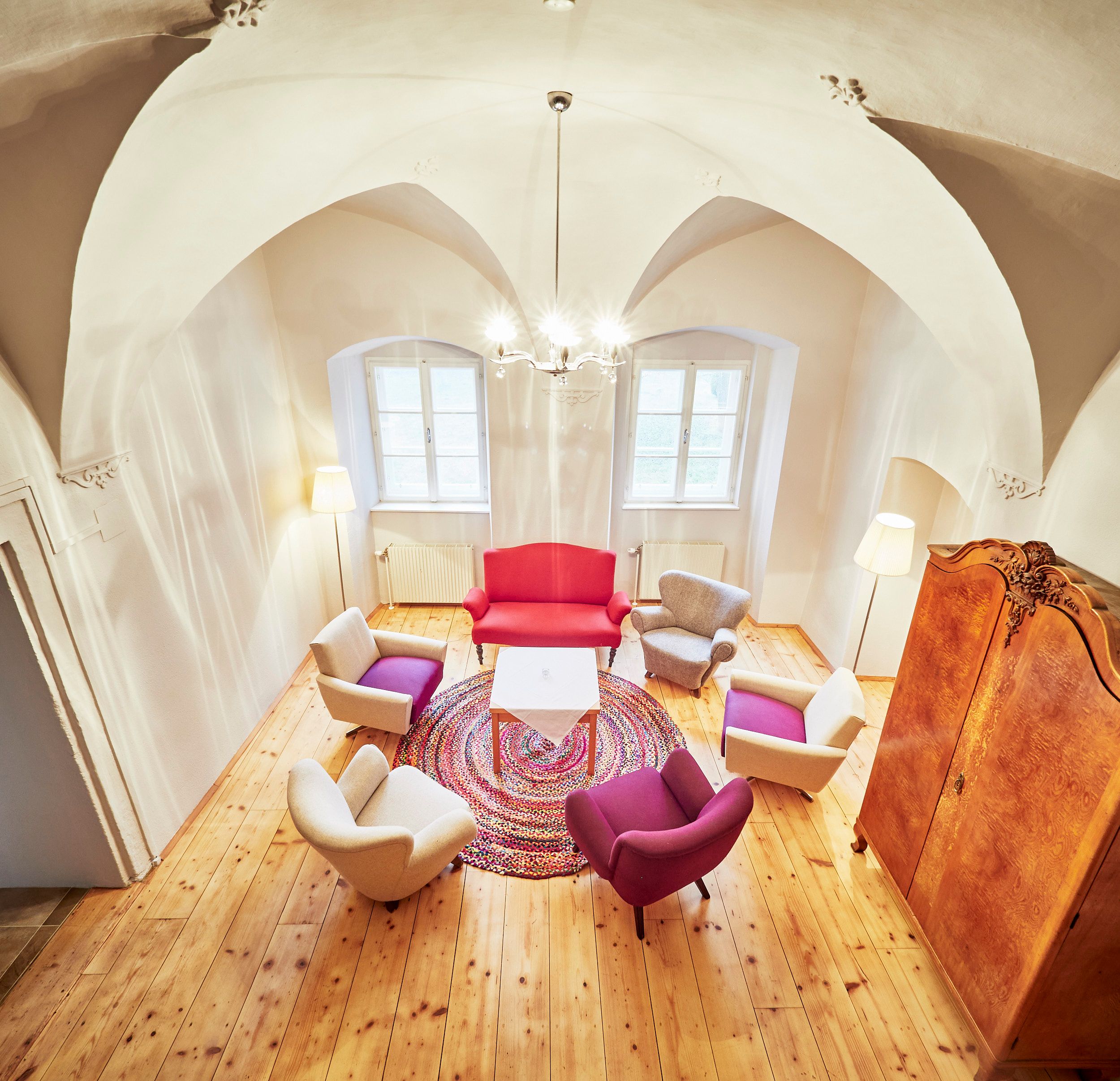 Ein gemütliches Teezimmer mit bunten Sesseln, einem roten Sofa und einem runden Teppich auf Holzboden. Ein Kronleuchter hängt von der gewölbten Decke.
