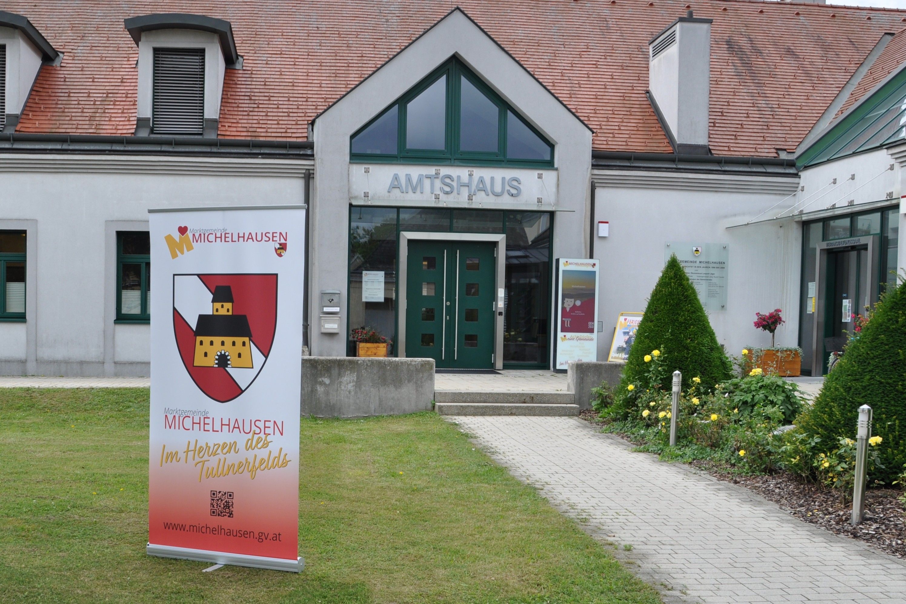 Eingang des Amtshauses Michelhausen mit Werbebanner und gepflegtem Garten.