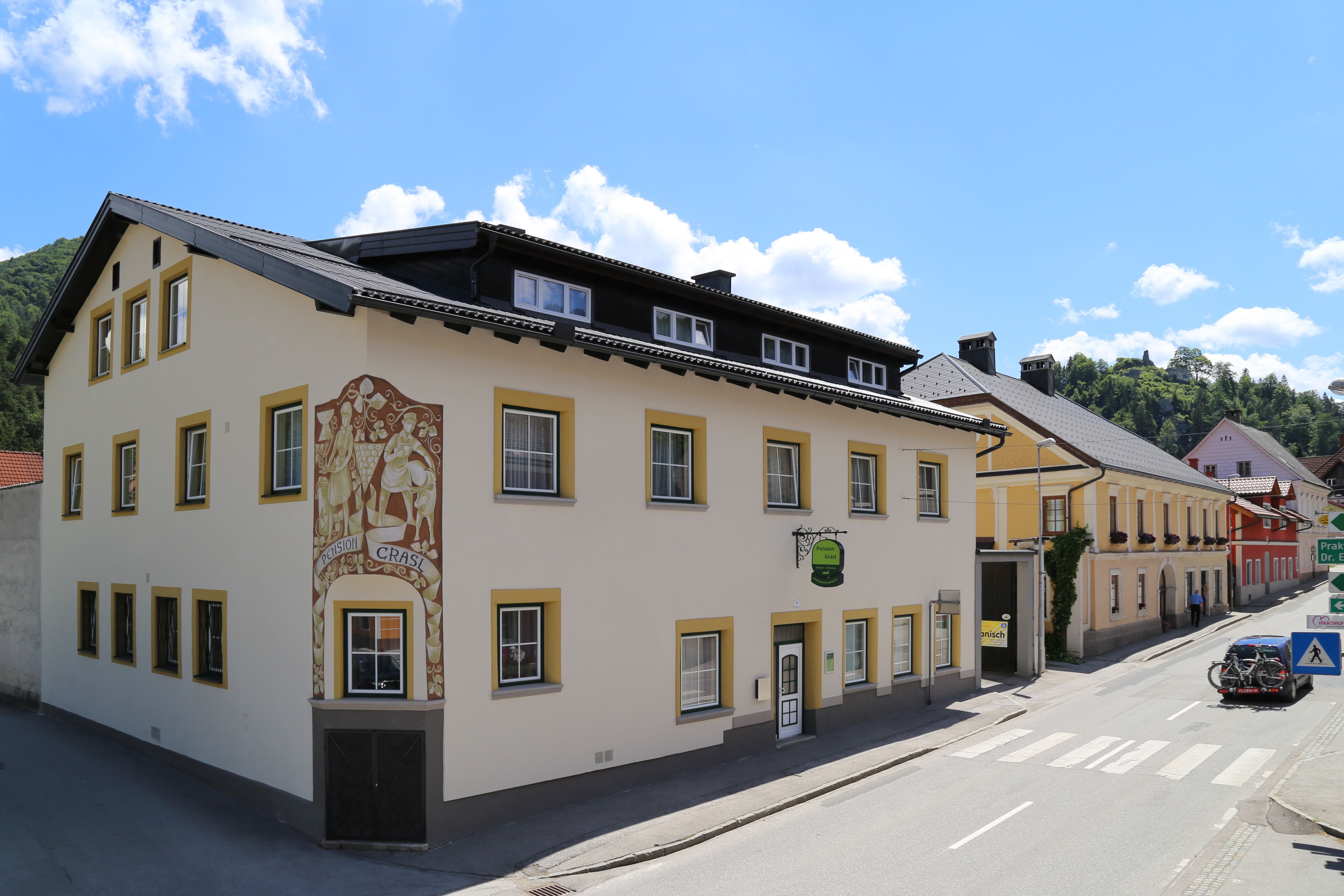 Ein gelbes Gebäude mit der Aufschrift 'Pension Grasl' an einer Straße bei sonnigem Wetter.