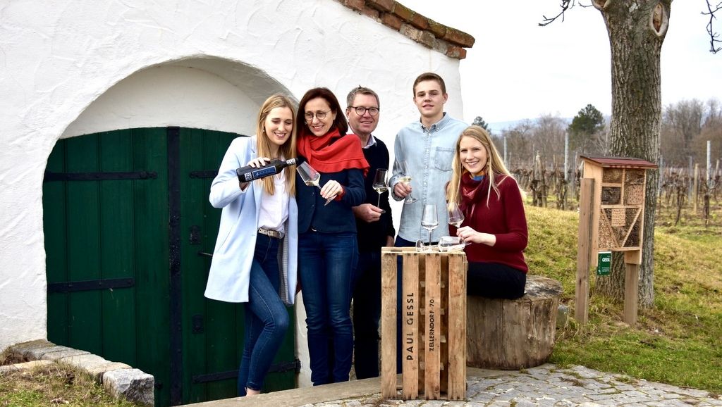 Eine Familie steht vor einem Weinkeller im Freien, mit Weingläsern und einer Weinflasche.