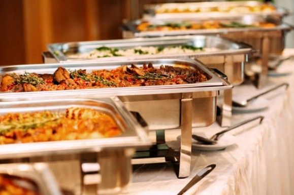 Buffet mit mehreren Edelstahl-Servierbehältern voller Speisen auf einem gedeckten Tisch.