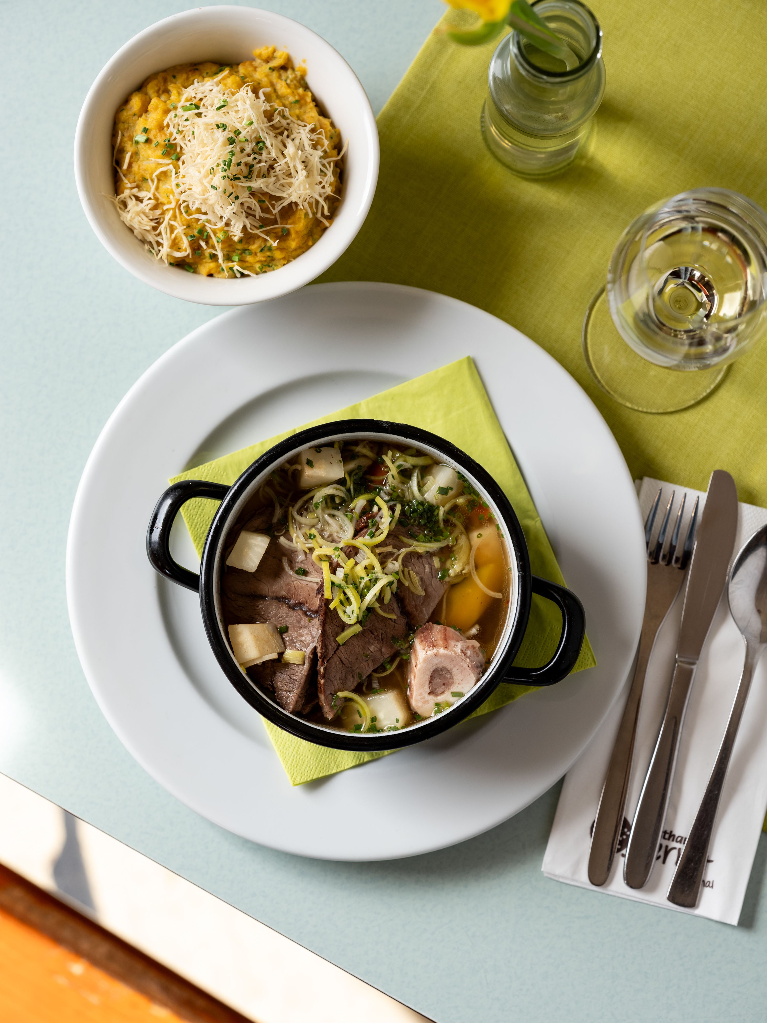 Ein Tisch mit einem Teller Suppe, einer Schüssel Brei mit Käse, einem Glas Wasser und Besteck auf einer grünen Serviette.