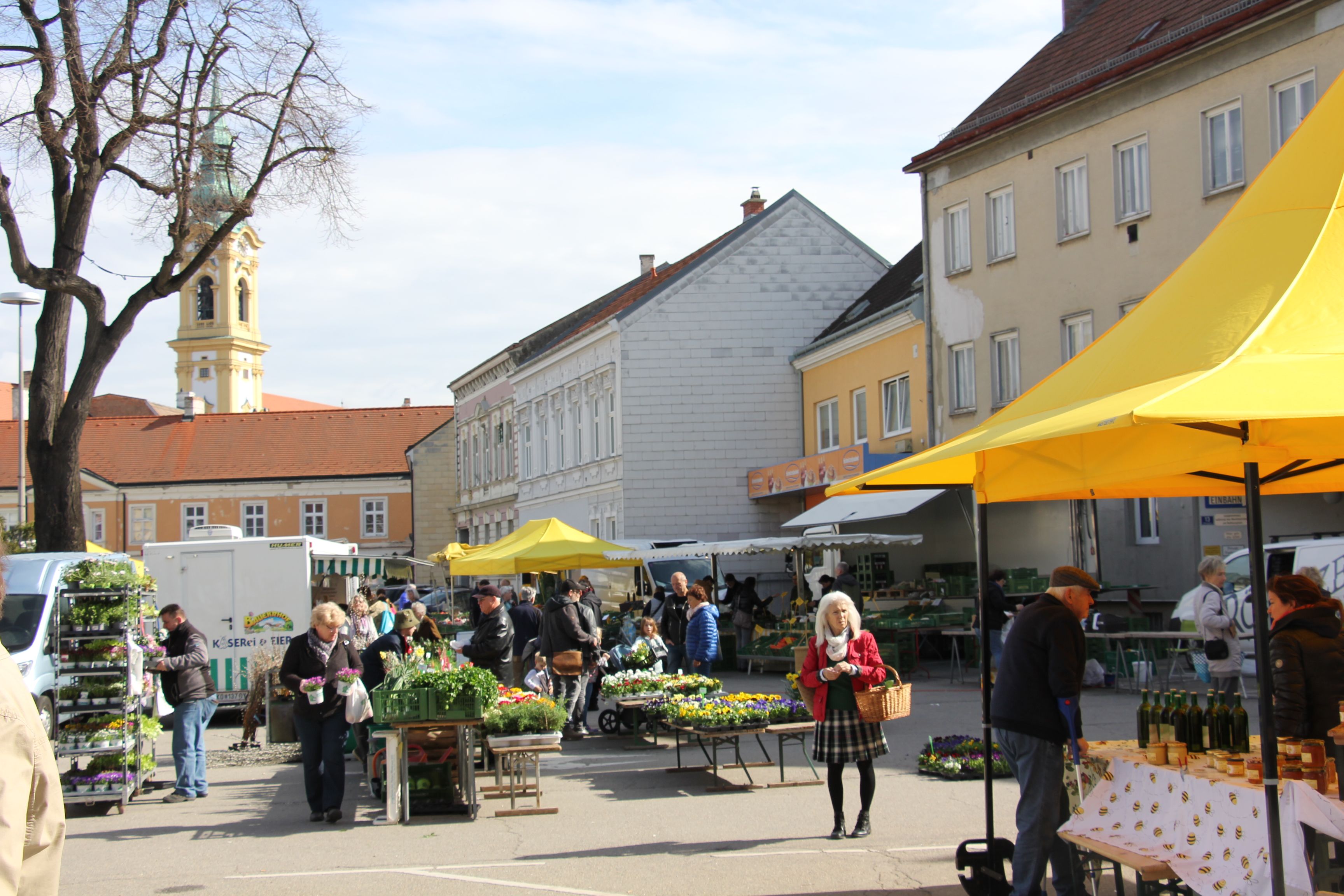 Menschen auf einem Wochenmarkt in Stockerau mit gelben Marktständen und einer Kirche im Hintergrund.