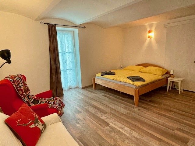 Gemütliches Schlafzimmer mit Holzboden, Doppelbett, rotem Sessel und Fenster mit Vorhang.