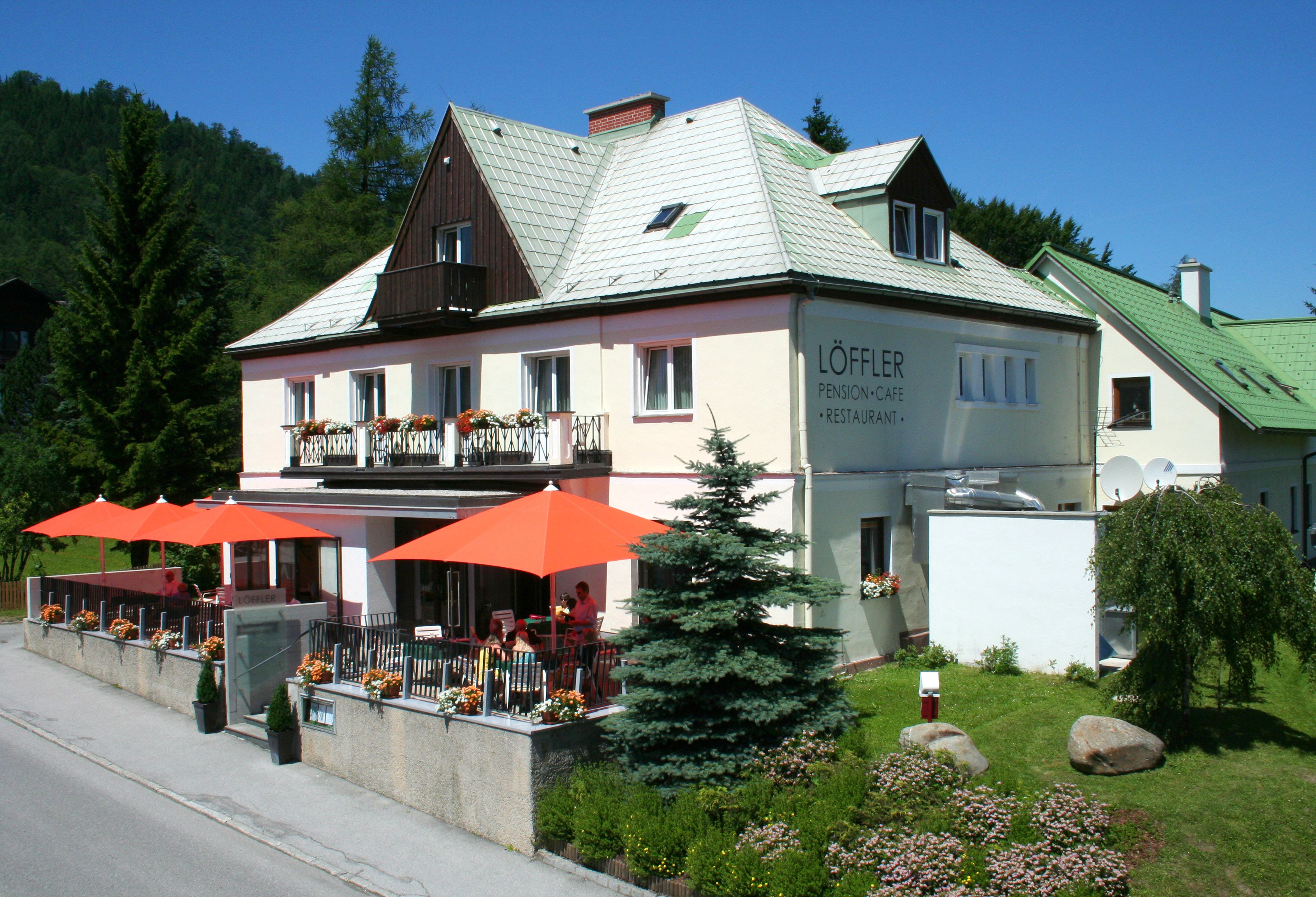 Ein zweistöckiges Gebäude mit der Aufschrift 'Löffler Pension-Café-Restaurant', umgeben von Bäumen und Blumen, mit orangenen Sonnenschirmen auf der Terrasse.