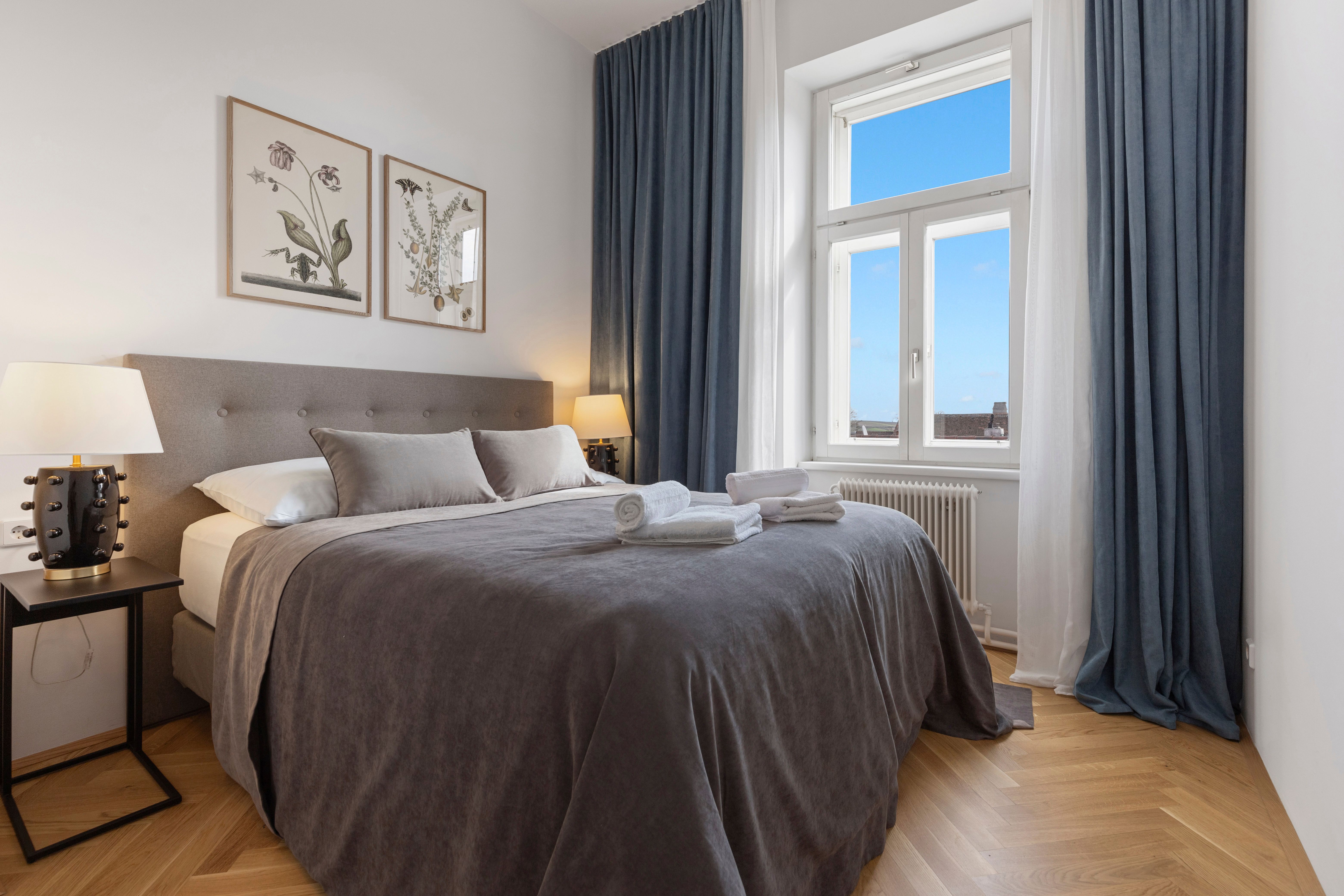Ein elegantes Schlafzimmer mit einem großen Bett, grauer Bettwäsche, zwei Nachttischen mit Lampen und einem Fenster mit blauem Vorhang.
