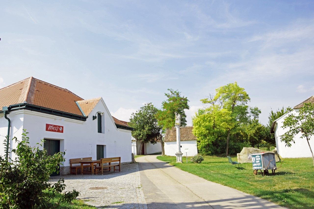 Kellergasse mit weißem Gebäude und Bänken vor einem Weingut an einem sonnigen Tag.