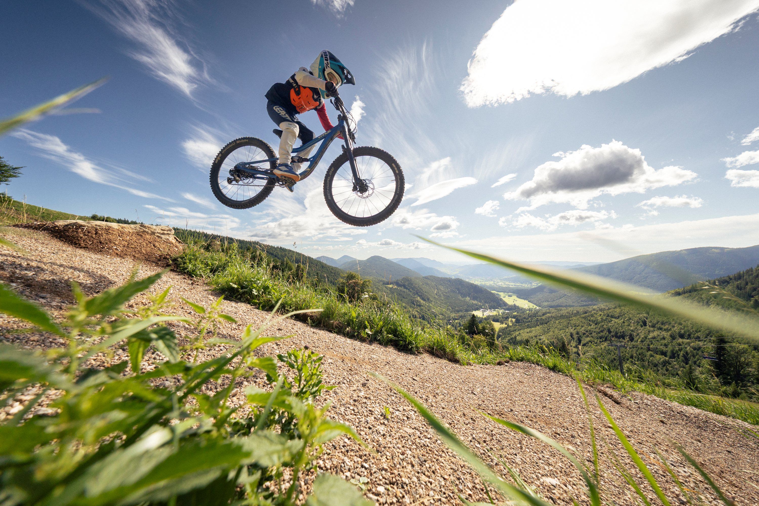 Ein Mountainbiker springt mit seinem Fahrrad über eine Rampe im Alpen Bikepark Schneeberg, umgeben von einer malerischen Berglandschaft unter blauem Himmel.