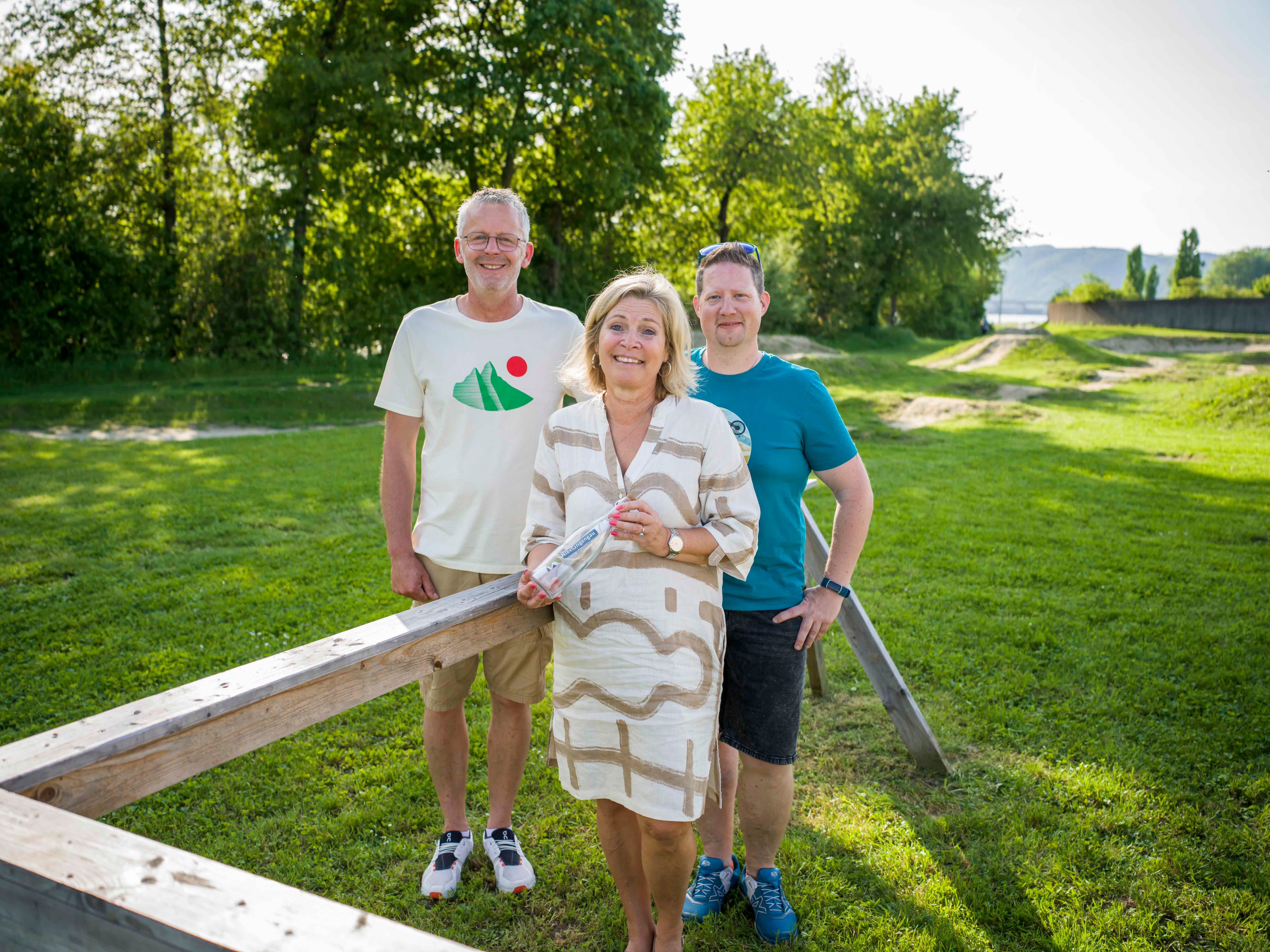 Drei Personen stehen auf einer Wiese vor einem Holzgeländer, umgeben von Bäumen.
