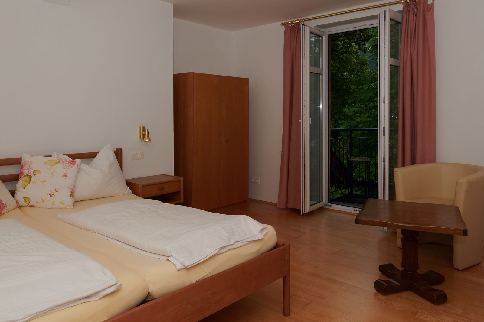 Schlafzimmer mit Doppelbett, Schrank, Tisch und Stuhl, Fenster mit Vorhängen.