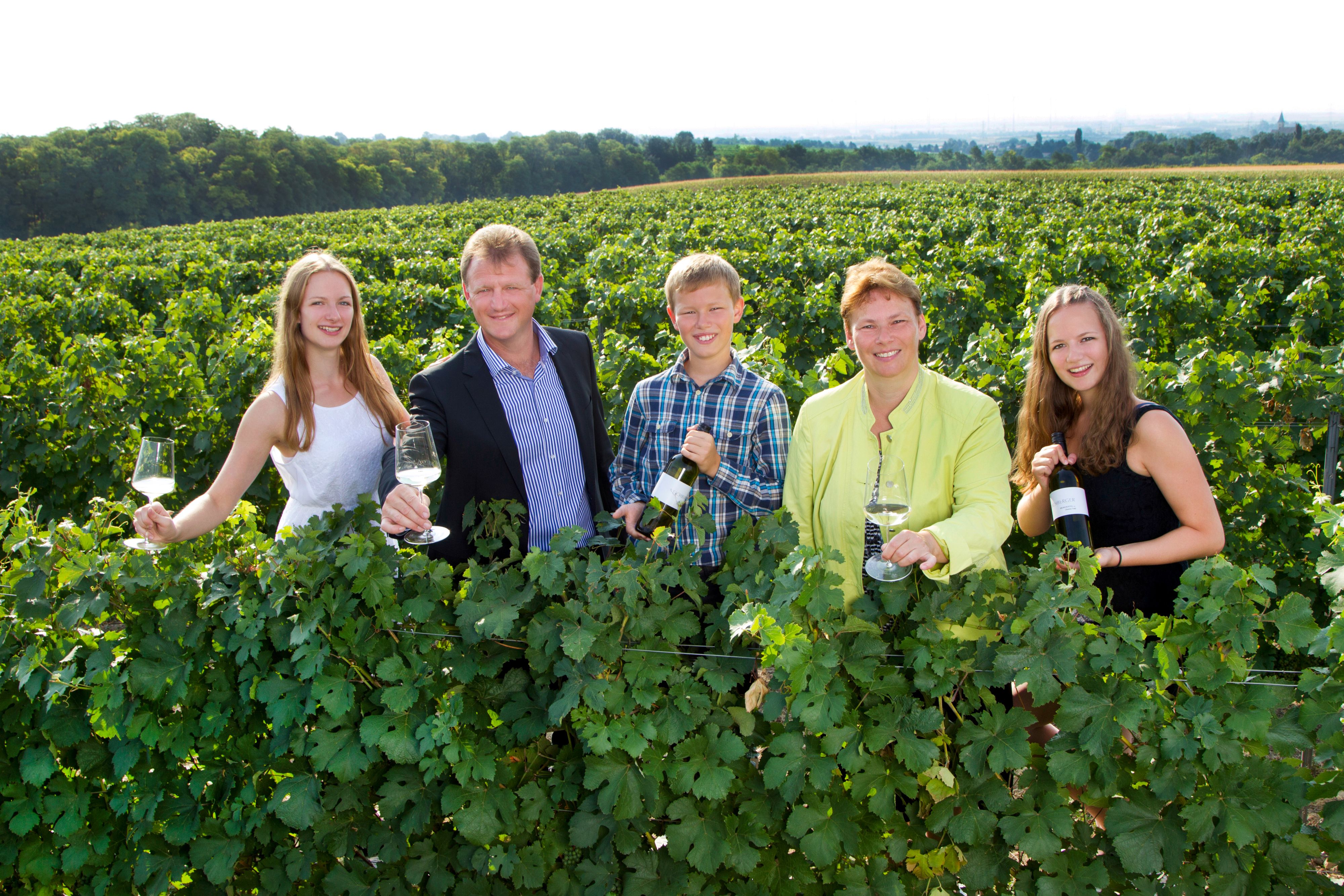 Eine Gruppe von fünf Personen steht in einem Weinberg und hält Weingläser und Flaschen.