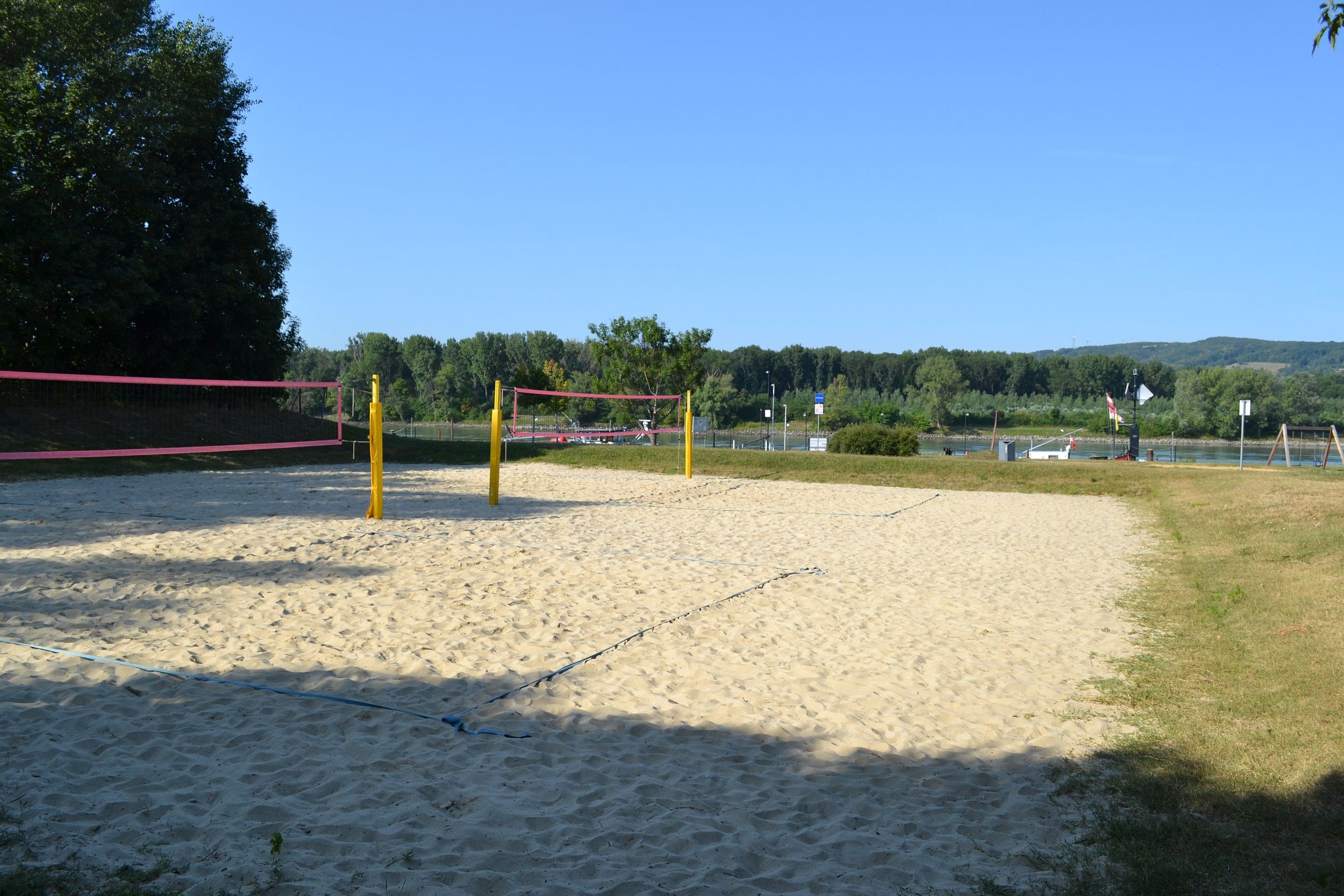 Ein leerer Beachvolleyballplatz mit gelben Pfosten und rosa Netz, umgeben von Bäumen und einem Fluss im Hintergrund.