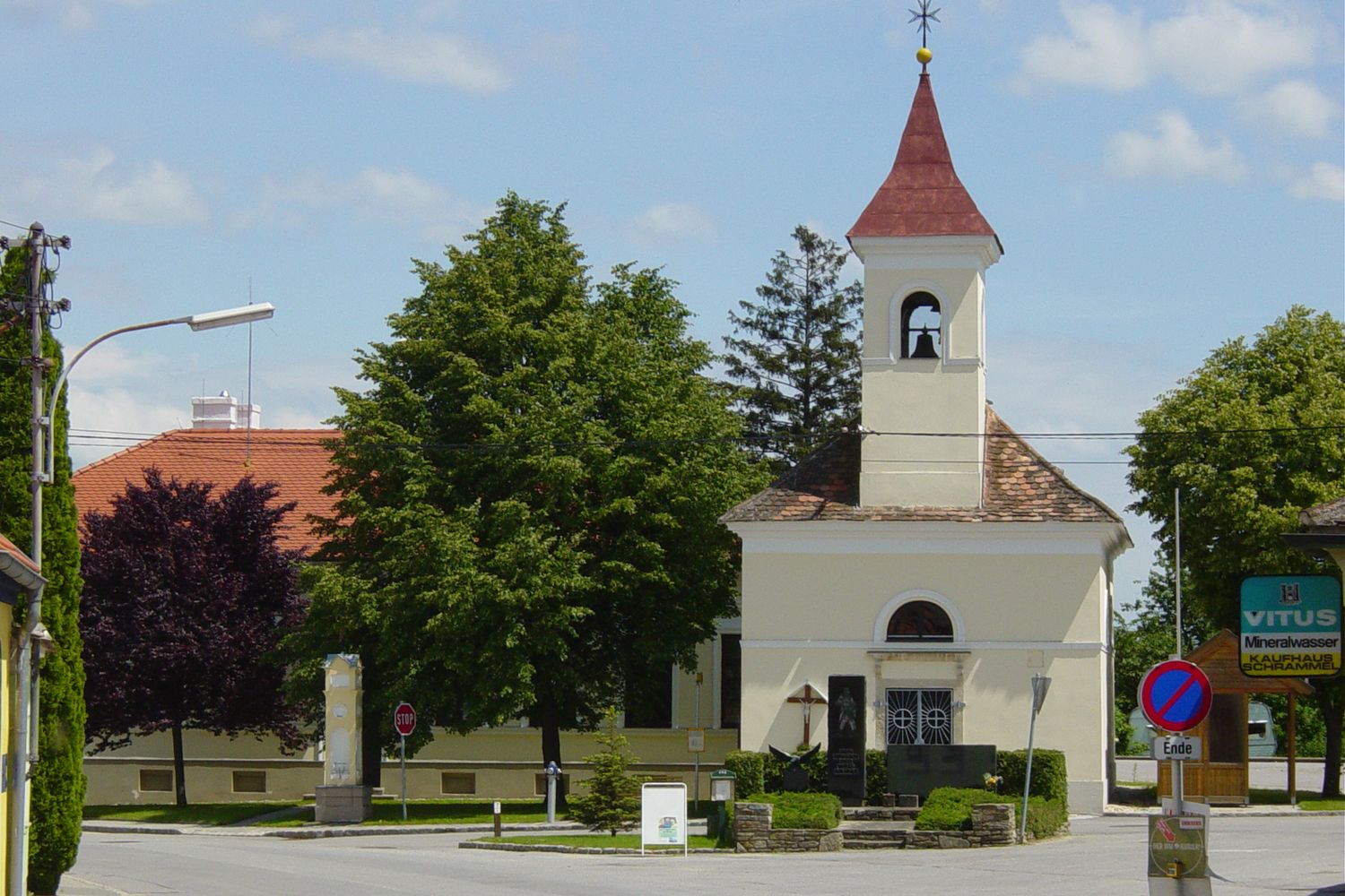 Kleine Kirche mit Glockenturm und rotem Dach, umgeben von Bäumen und Straßenschildern.