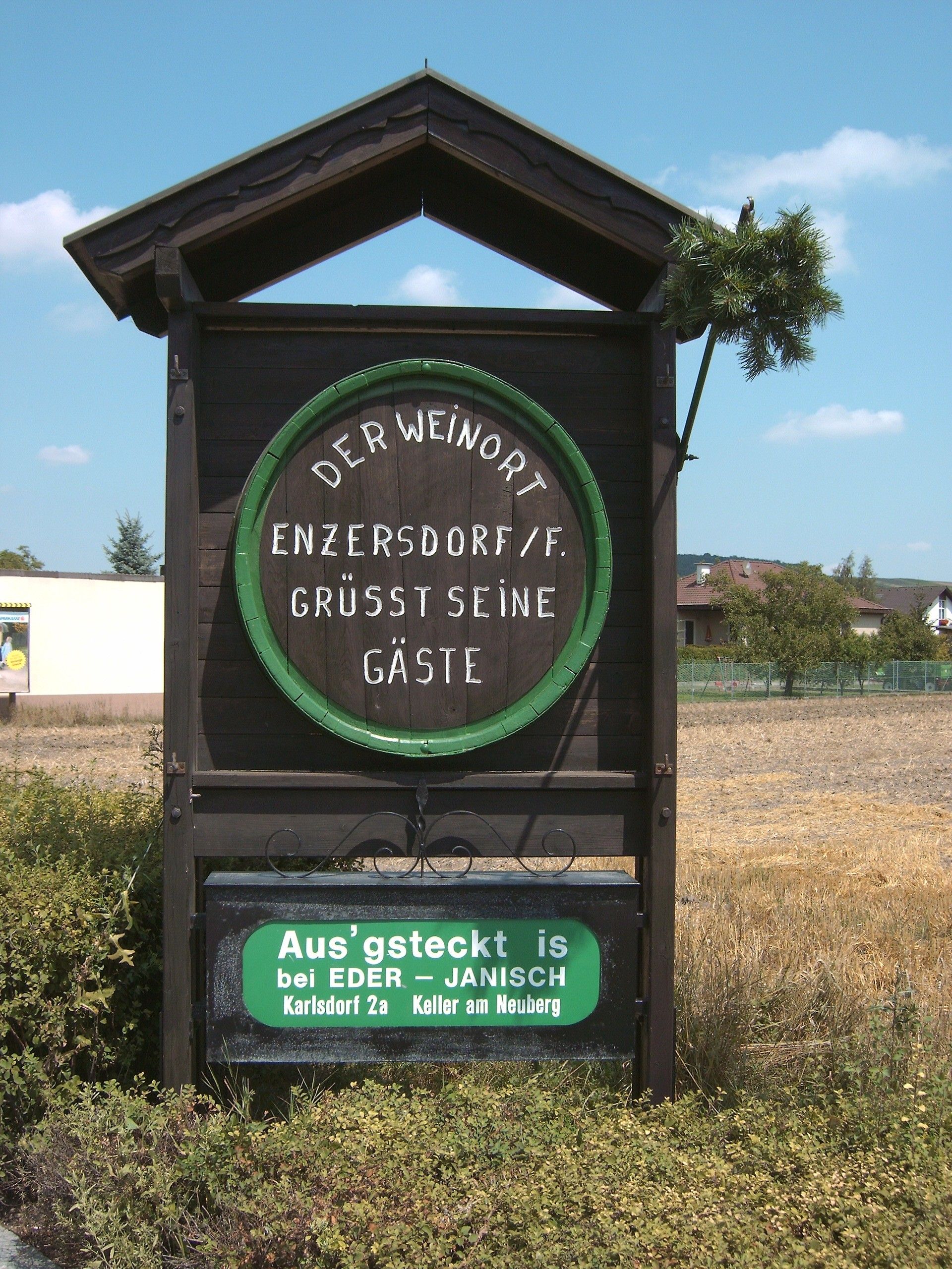 Holztafel in Enzersdorf an der Fischa mit Begrüßungstext und Hinweis auf Heurigenbetrieb.