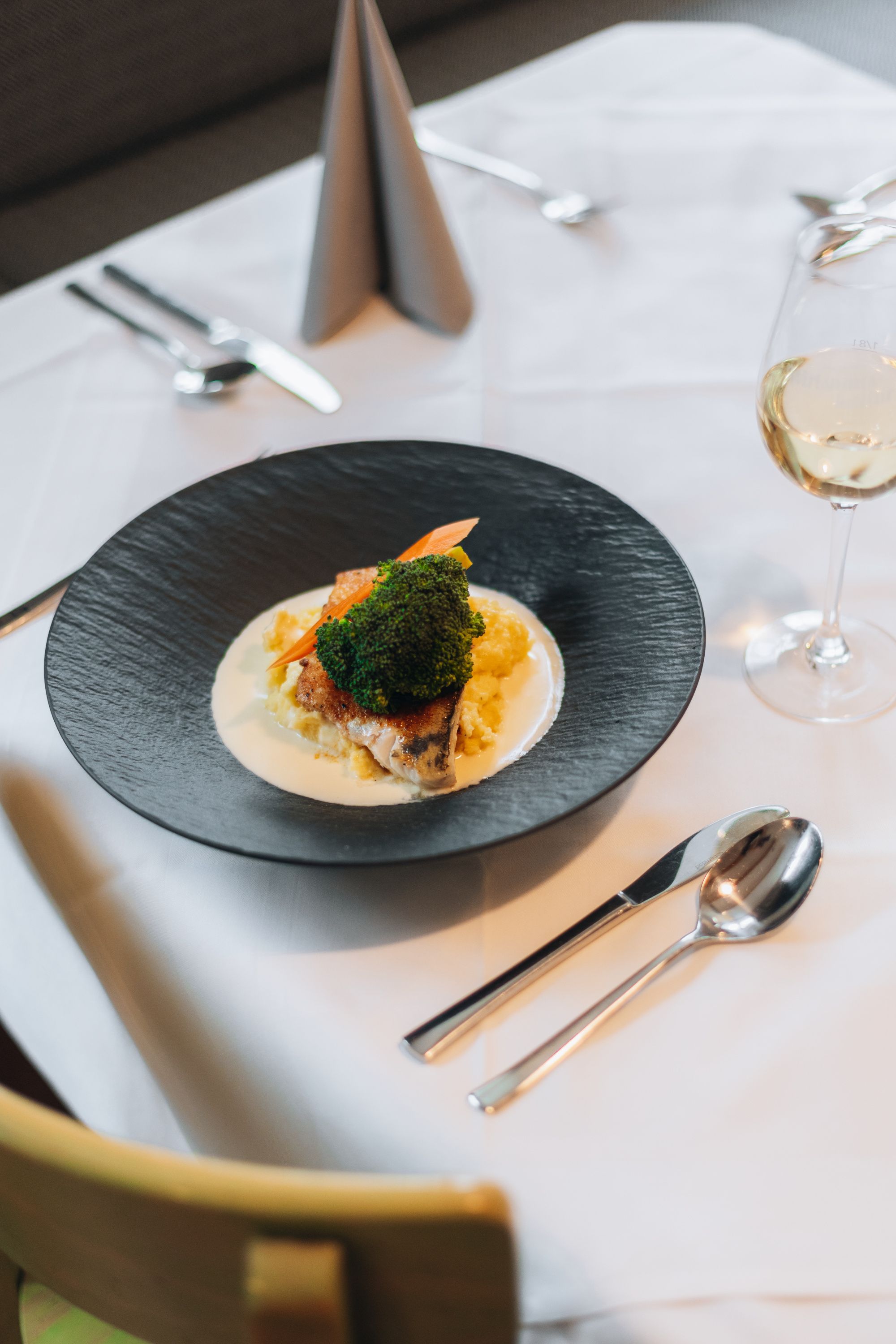 Ein elegant angerichtetes Gericht auf einem schwarzen Teller mit Gemüse und Sauce, daneben ein Glas Weißwein auf einem gedeckten Tisch.