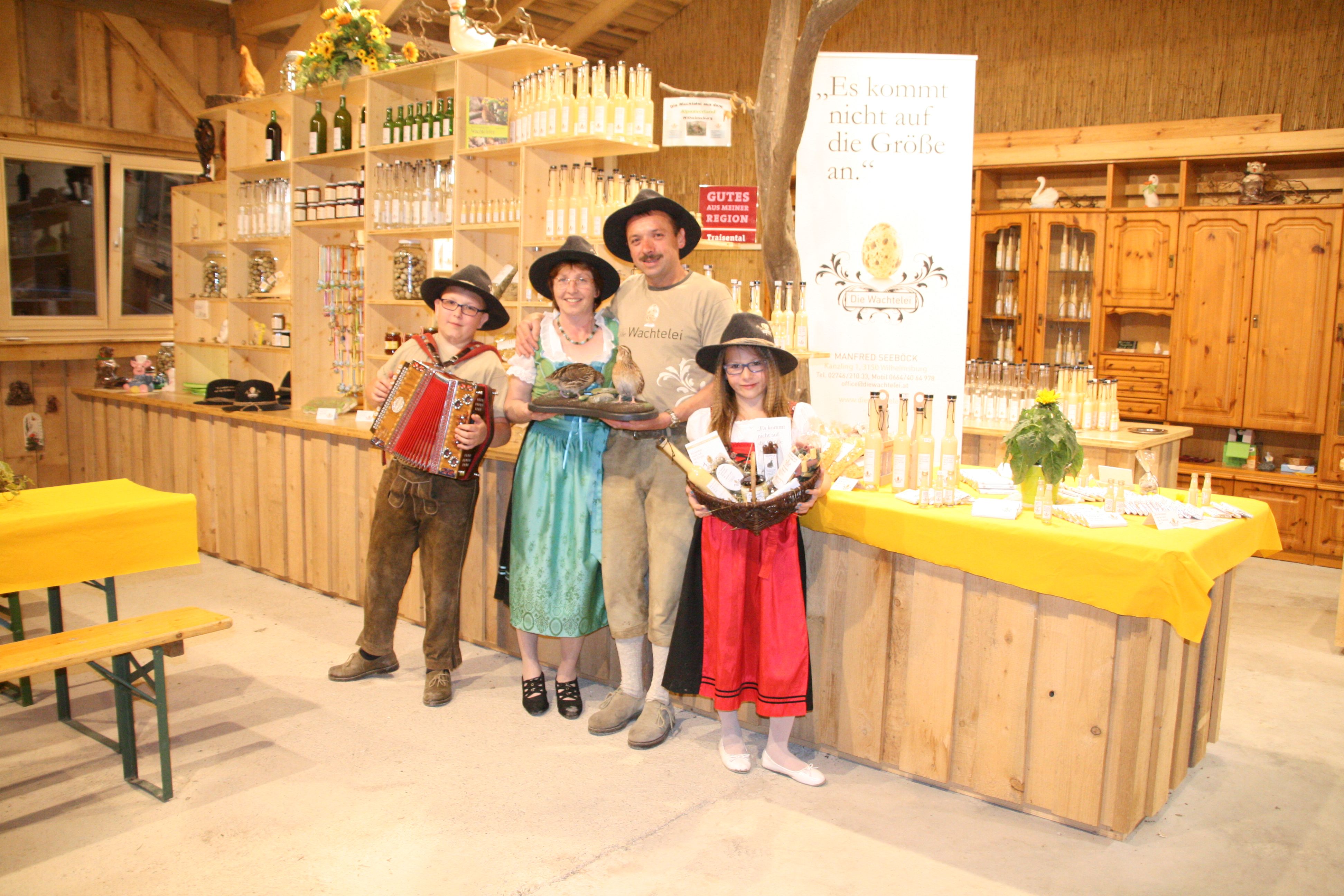 Eine Familie in traditioneller Kleidung steht in einem Laden mit Produkten und einem Banner mit der Aufschrift 'Die Wachtelei'.